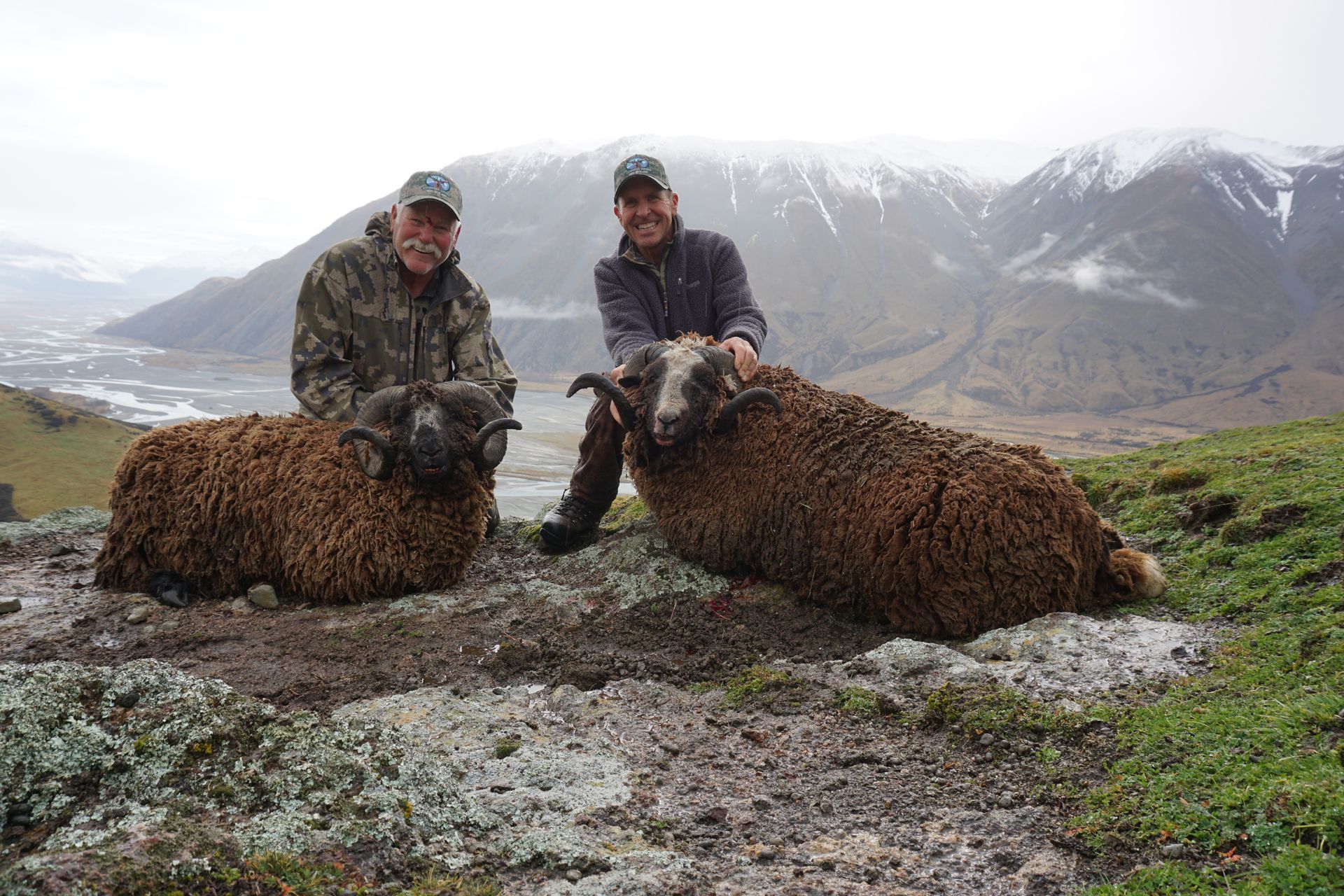 Two men pose with two large sheep with curled horns, against a mountain backdrop on a cloudy day.