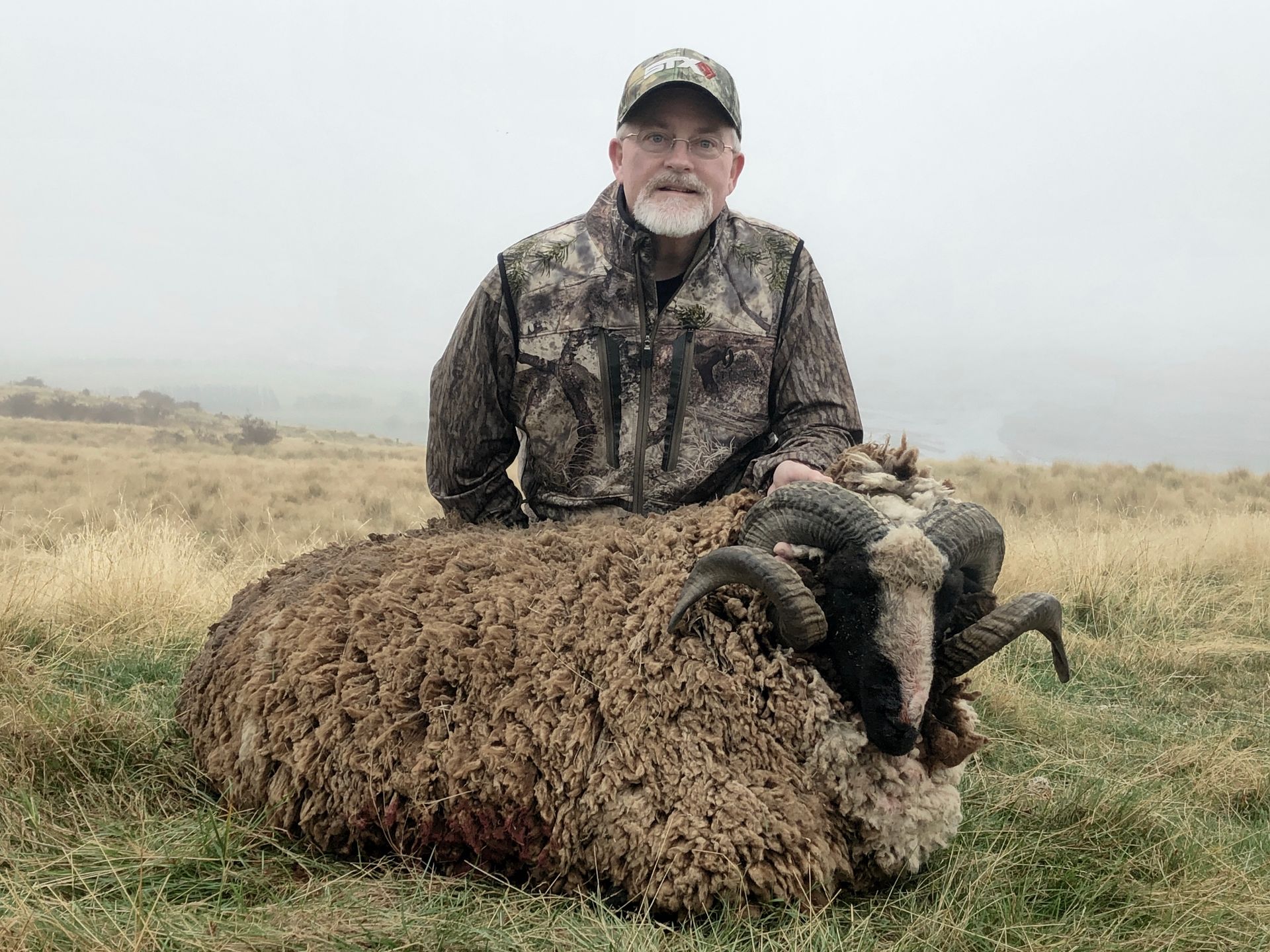 Man in camouflage gear poses with a large, dead ram in a grassy field, overcast sky.