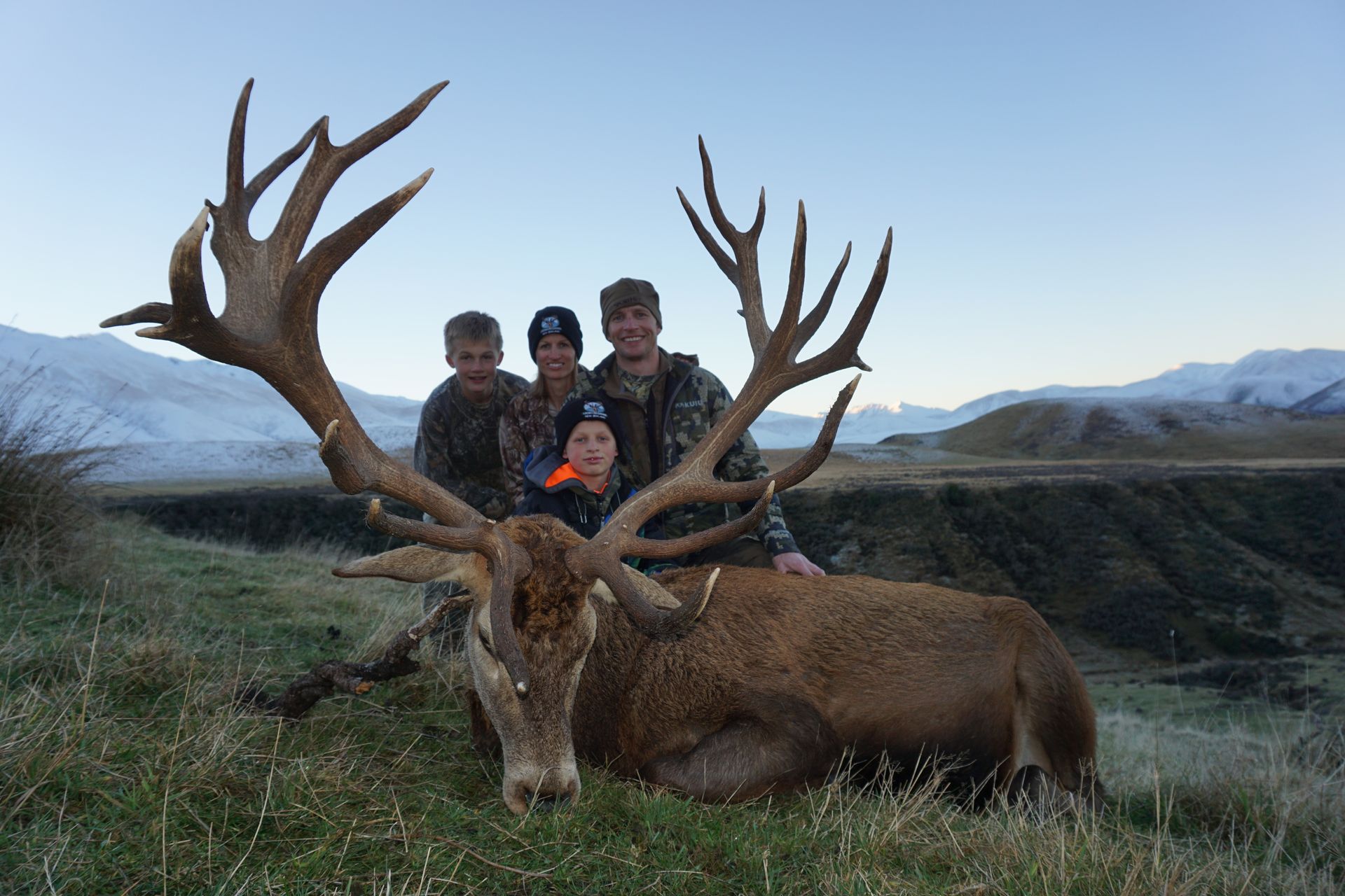 Four people posing with a large dead red stag in a grassy field, mountains in the background. The stag has large antlers, and the people are smiling.