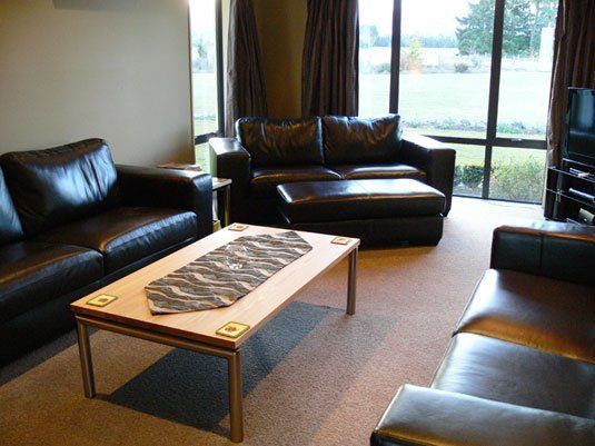 Living room with black leather furniture, a light wood coffee table, and a view through large windows.
