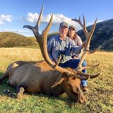 Man and woman pose next to a large elk lying on grass. The man sits on the elk; rolling hills are in the background.