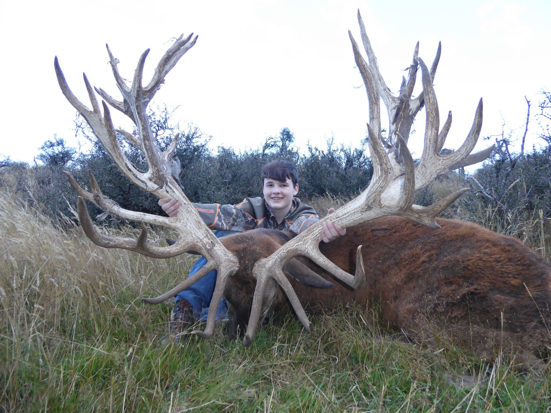 Boy in camouflage holds up the large antlers of a dead red deer stag in a grassy field.