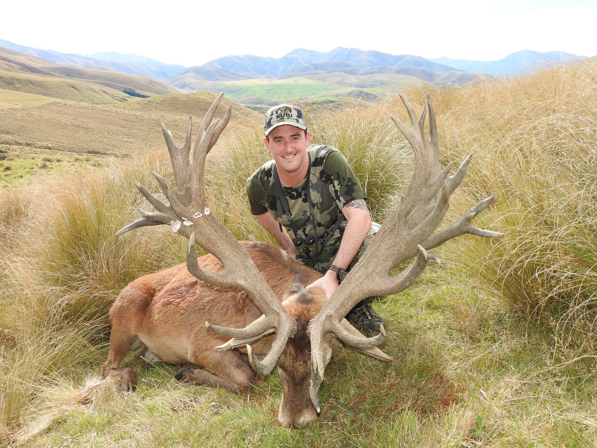 Man kneels next to a large, dead red deer buck with impressive antlers, in a grassy mountain setting. The man smiles.