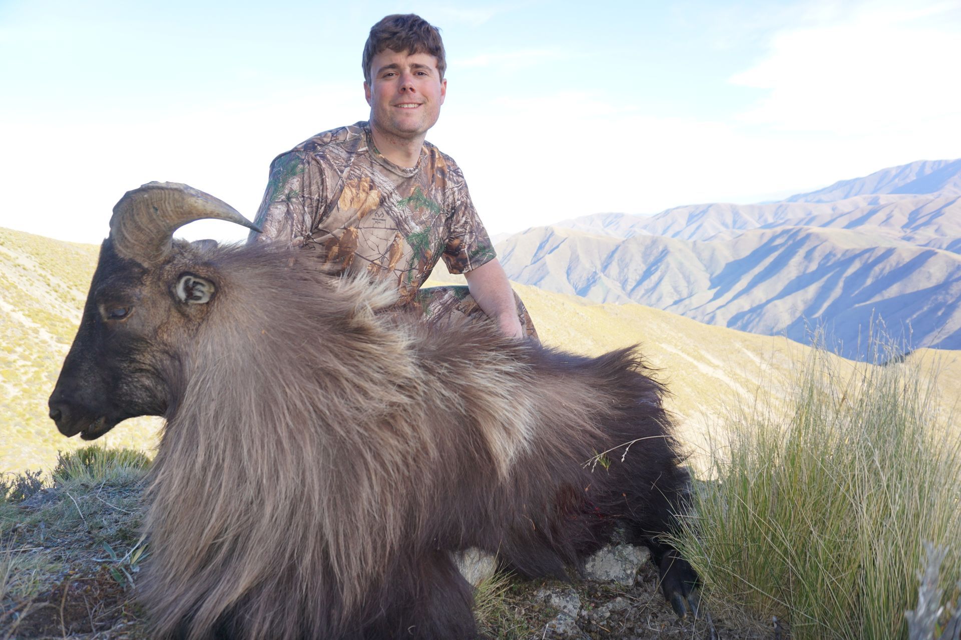 Man in camouflage posing with a dead Himalayan tahr in a mountainous landscape. The tahr has large horns and brown fur.
