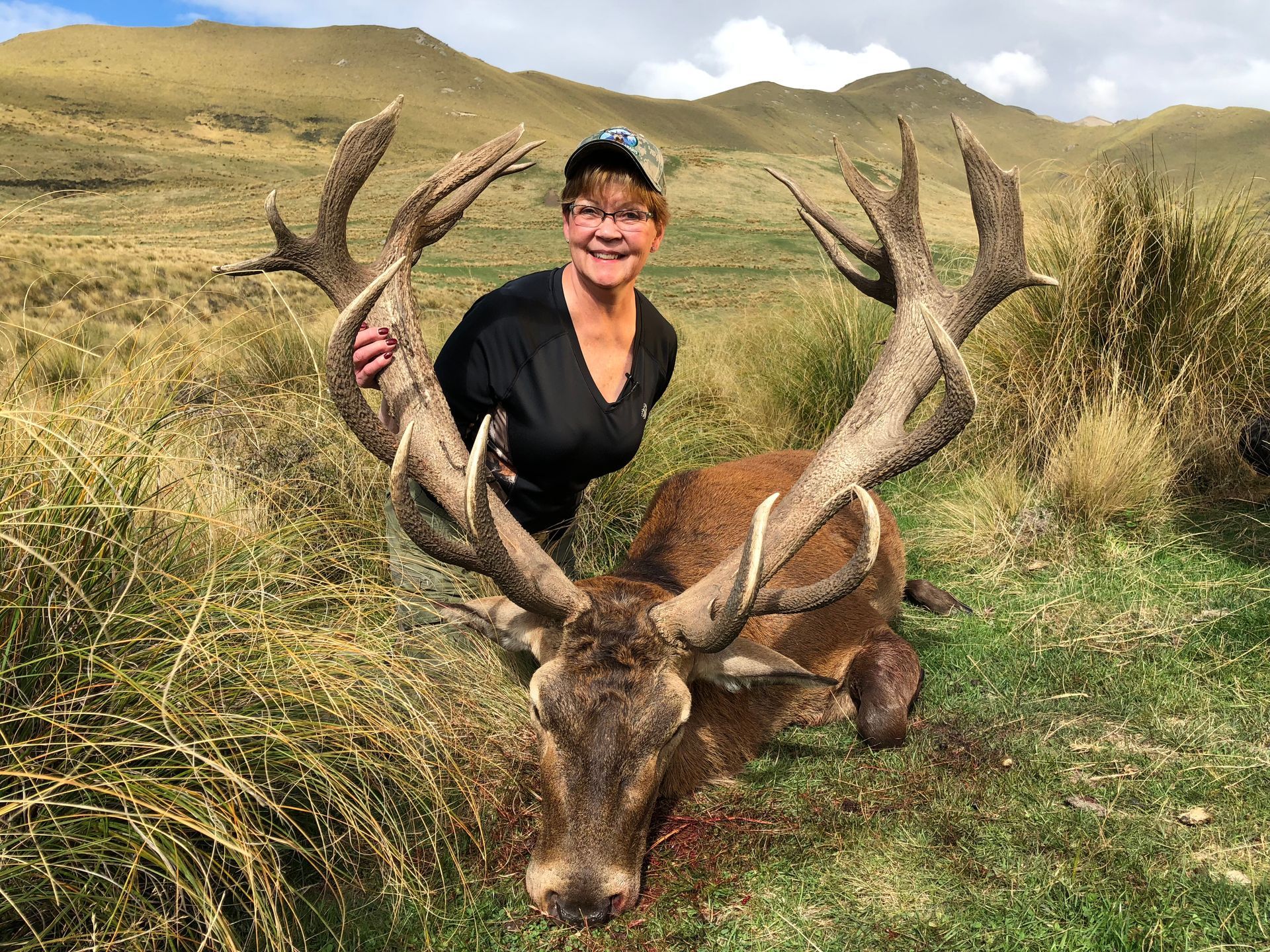 Woman in a camouflage hat smiles, kneeling next to a large dead stag with impressive antlers in a grassy field.