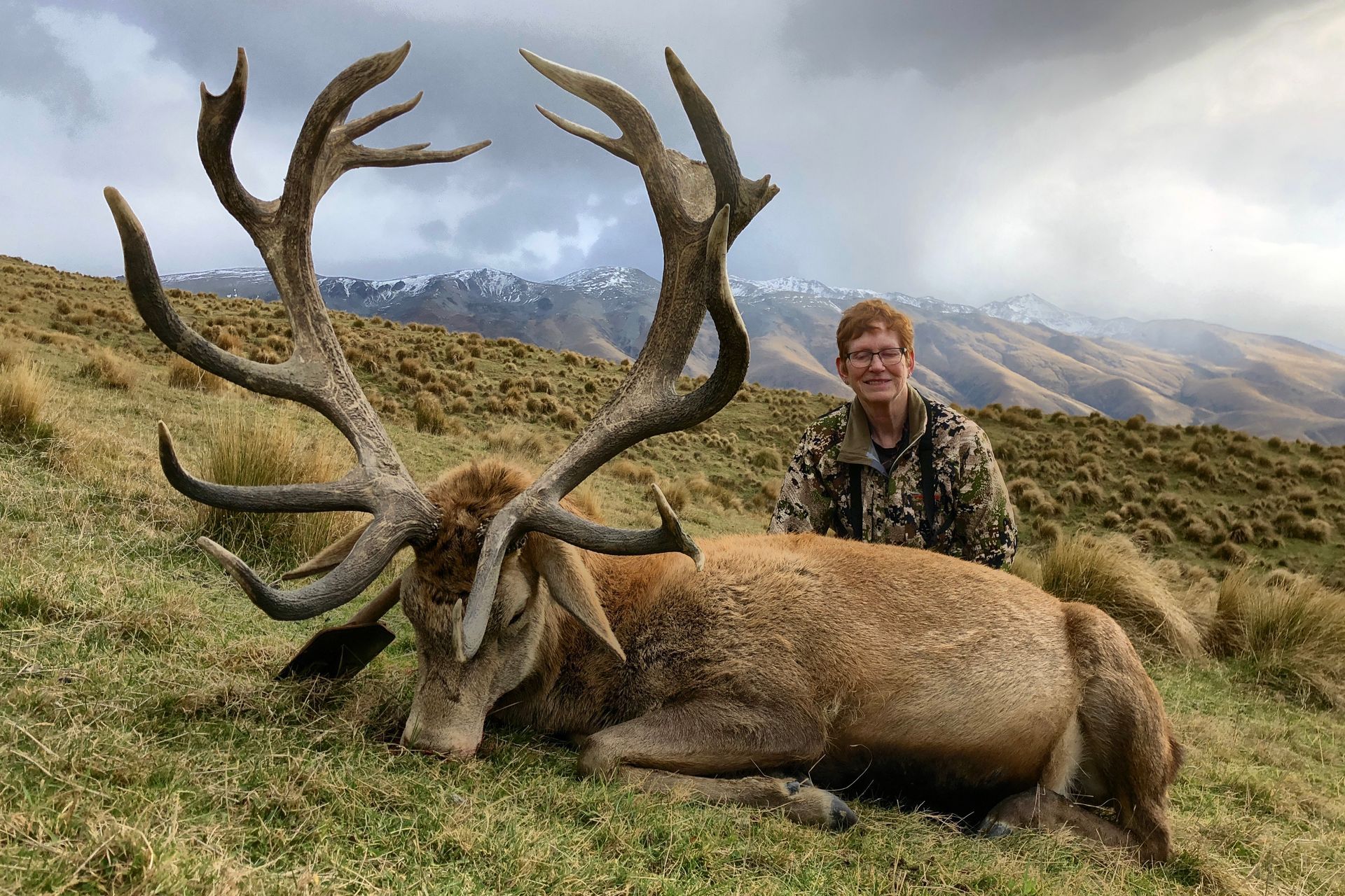 Hunter in camouflage with a large red deer. Deer lies on grass with large antlers; mountains in the background.