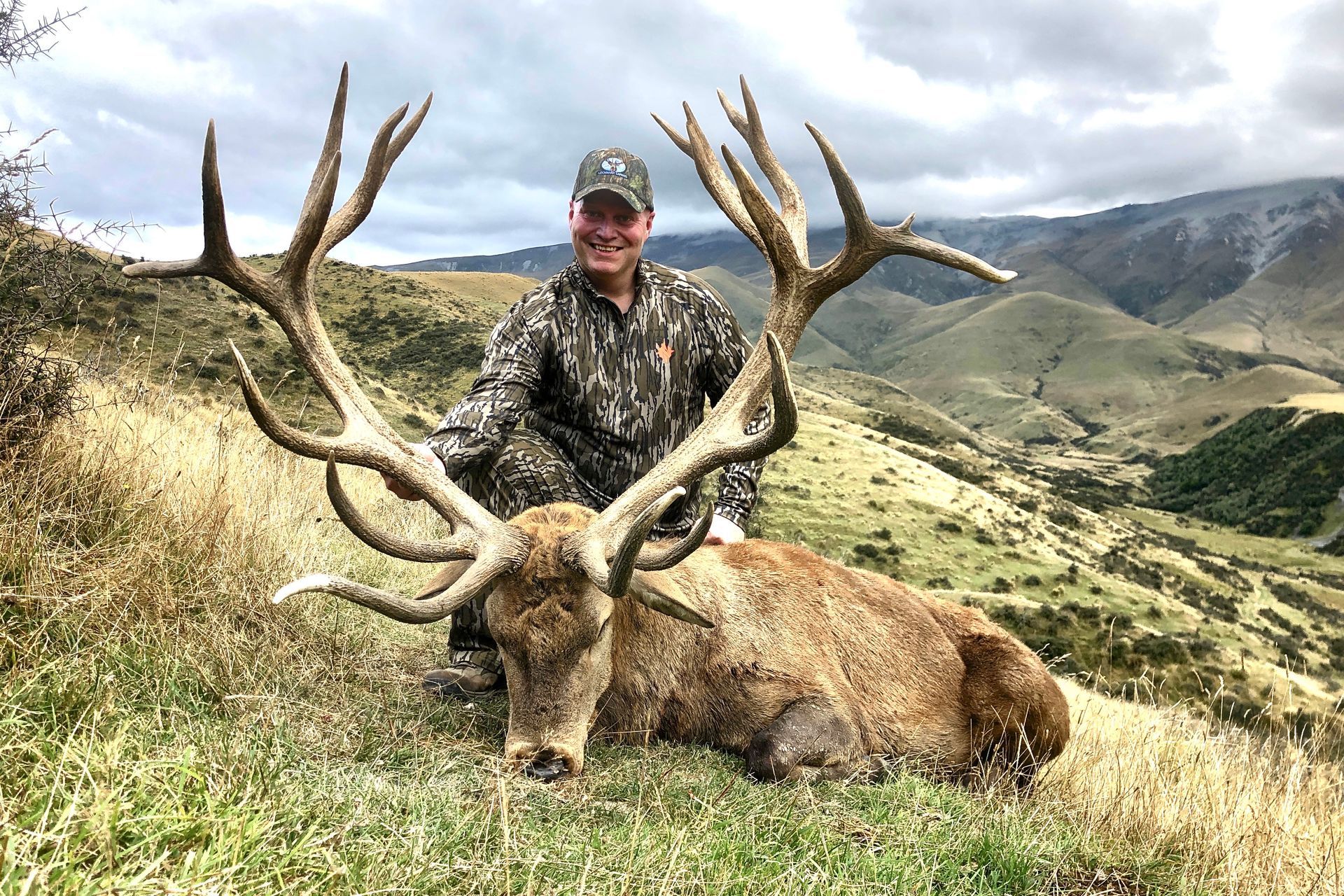 Man in camouflage kneeling next to a large dead red deer with impressive antlers in a grassy mountain landscape.