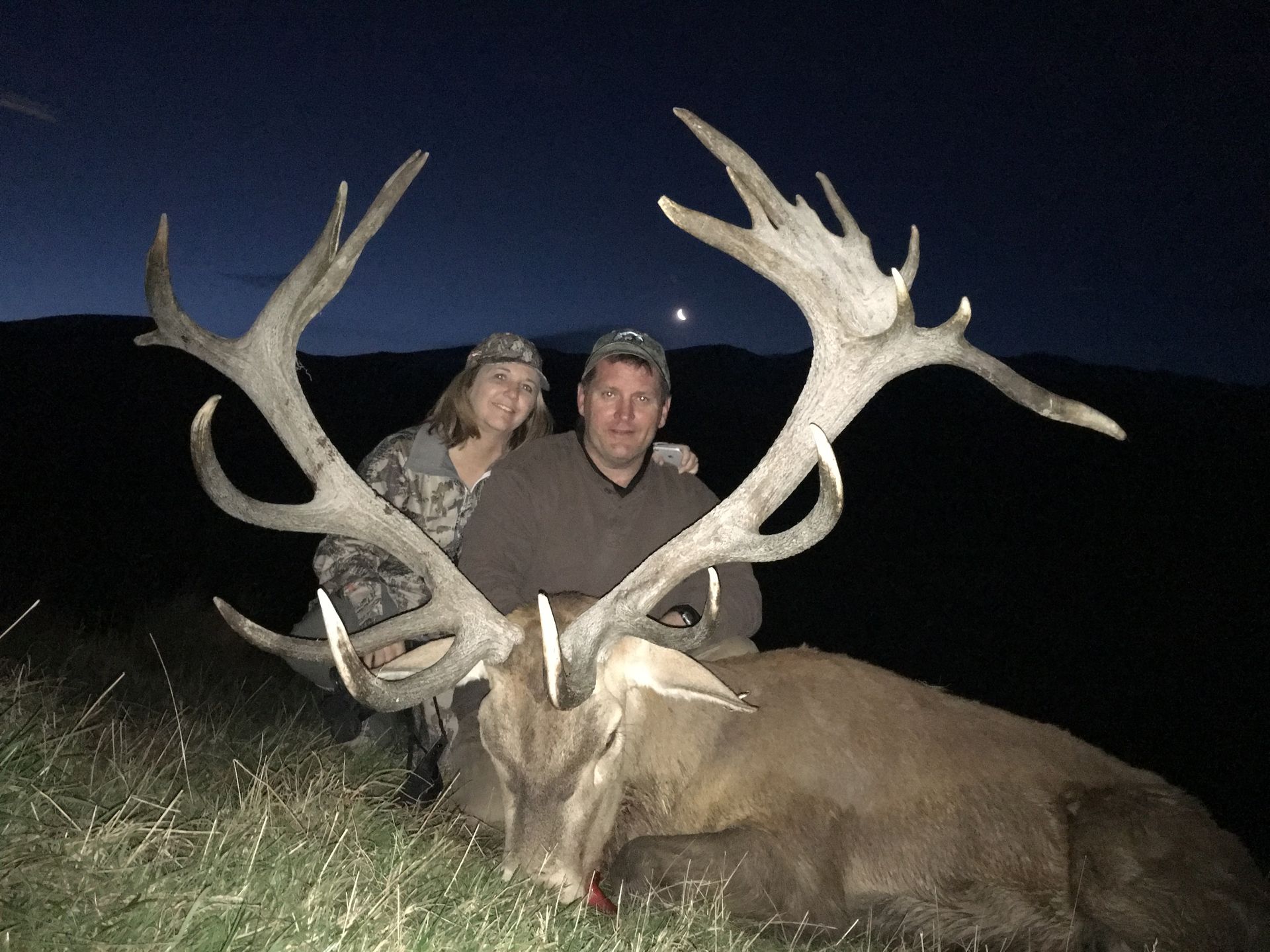 A man and woman pose with a large deer displaying massive antlers at dusk. They are outdoors in a grassy area.