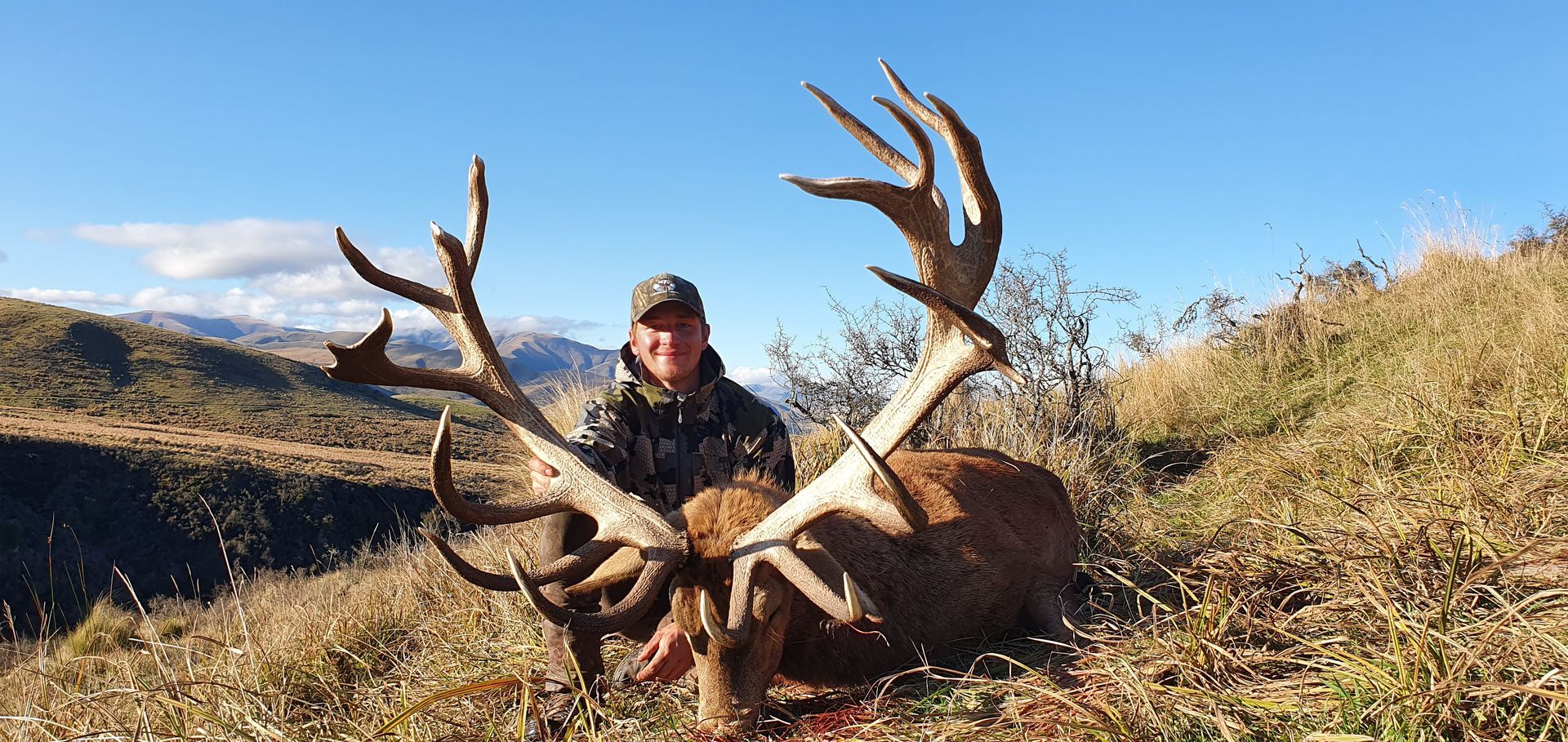 A man in camouflage holds a large dead elk with impressive antlers, posed in a grassy outdoor setting under a blue sky.