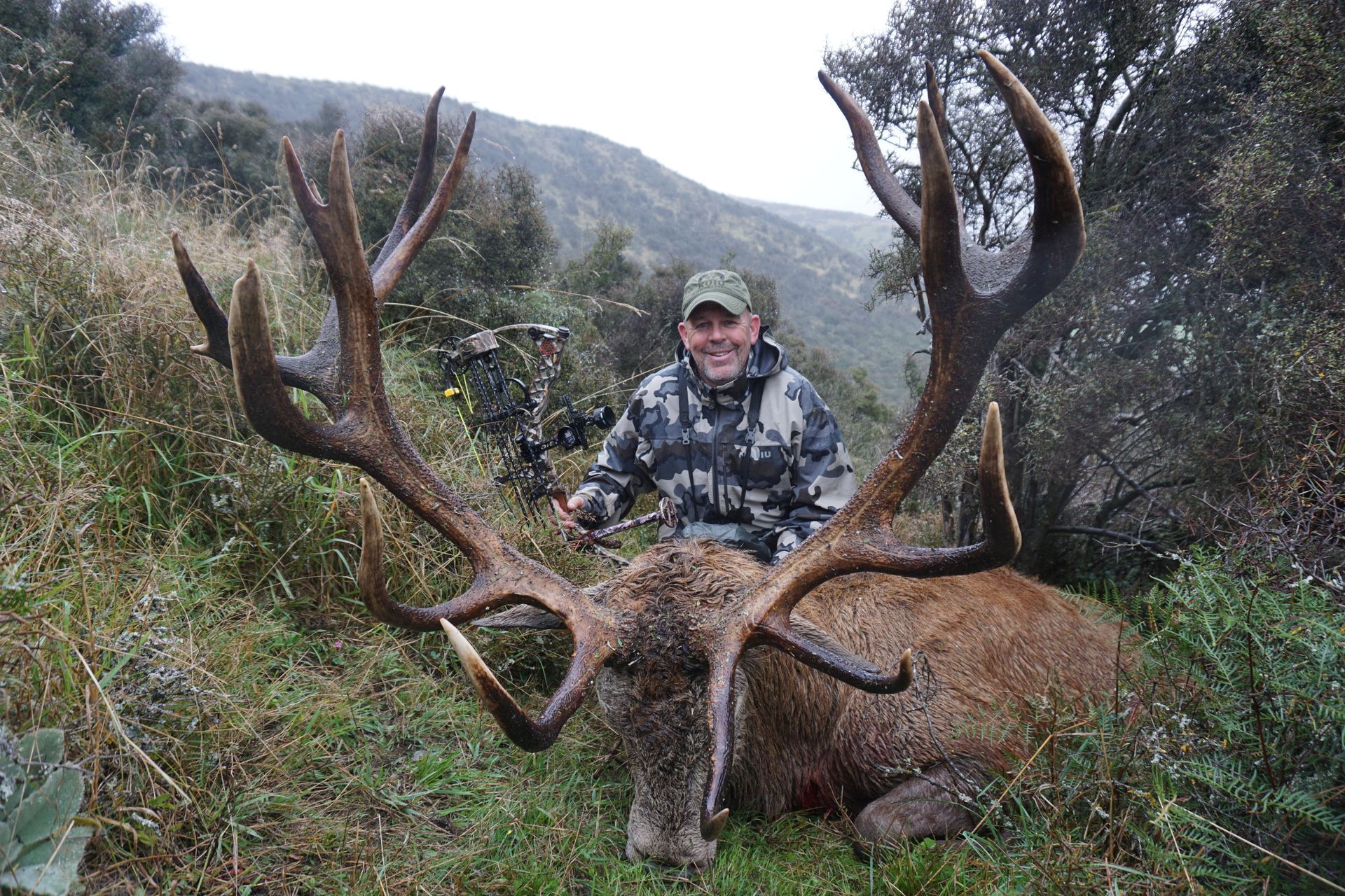 Man in camouflage smiles beside a large red deer he hunted, with large antlers in a grassy hillside setting.