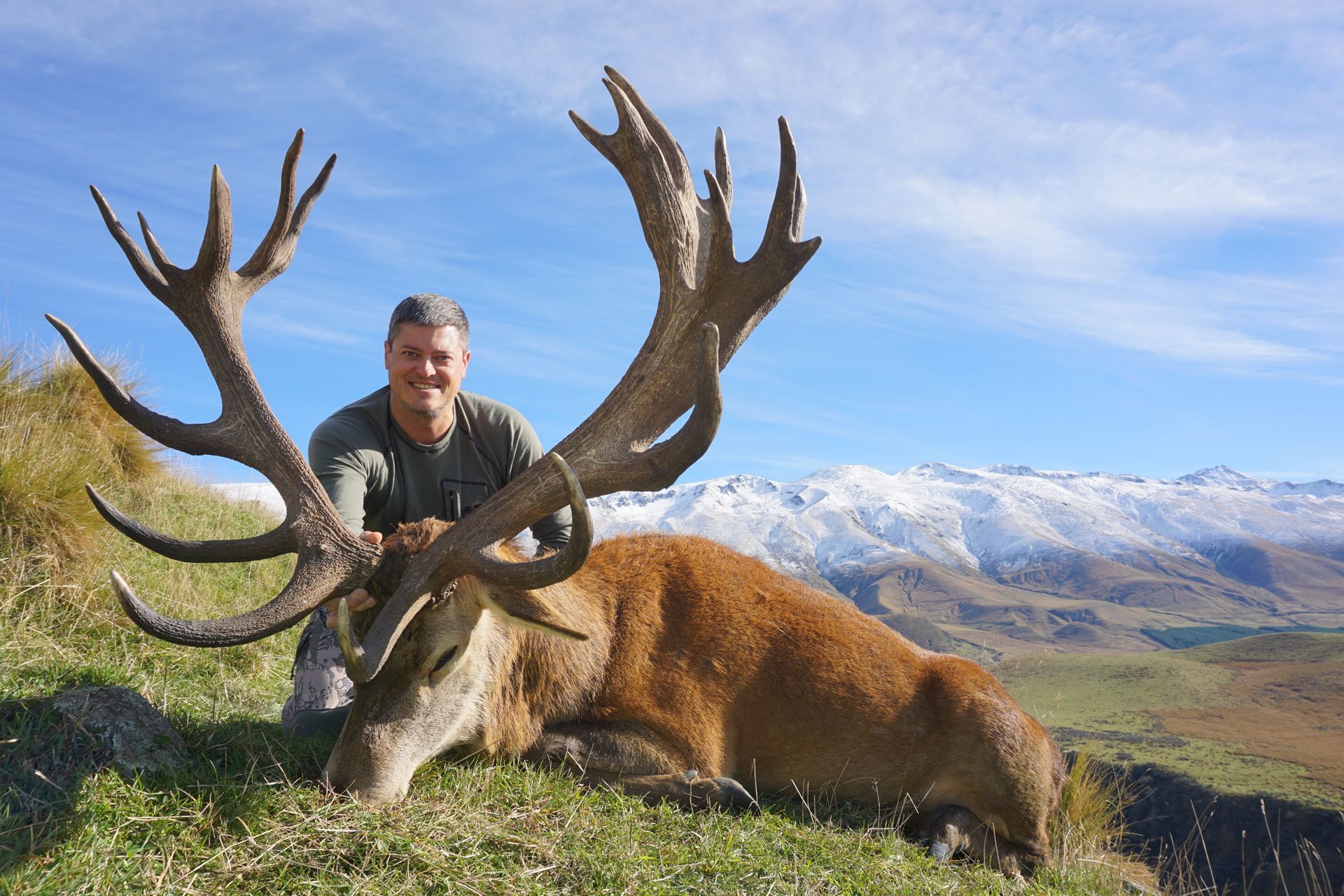 Man kneels beside a large, reddish-brown stag with massive antlers on a grassy hillside with mountains in the background.
