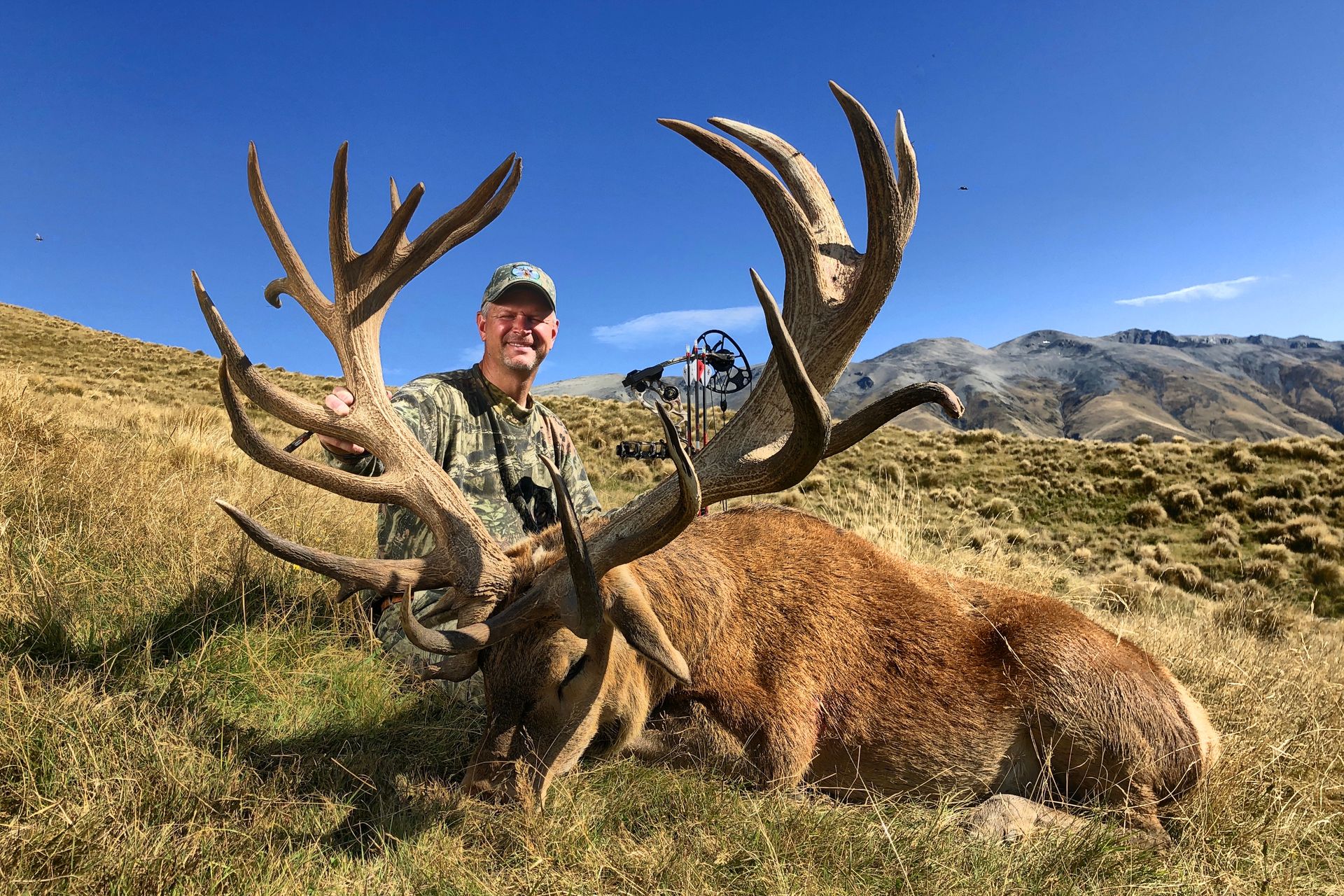 Man in camouflage kneeling next to a large dead red deer with enormous antlers in a grassy field.