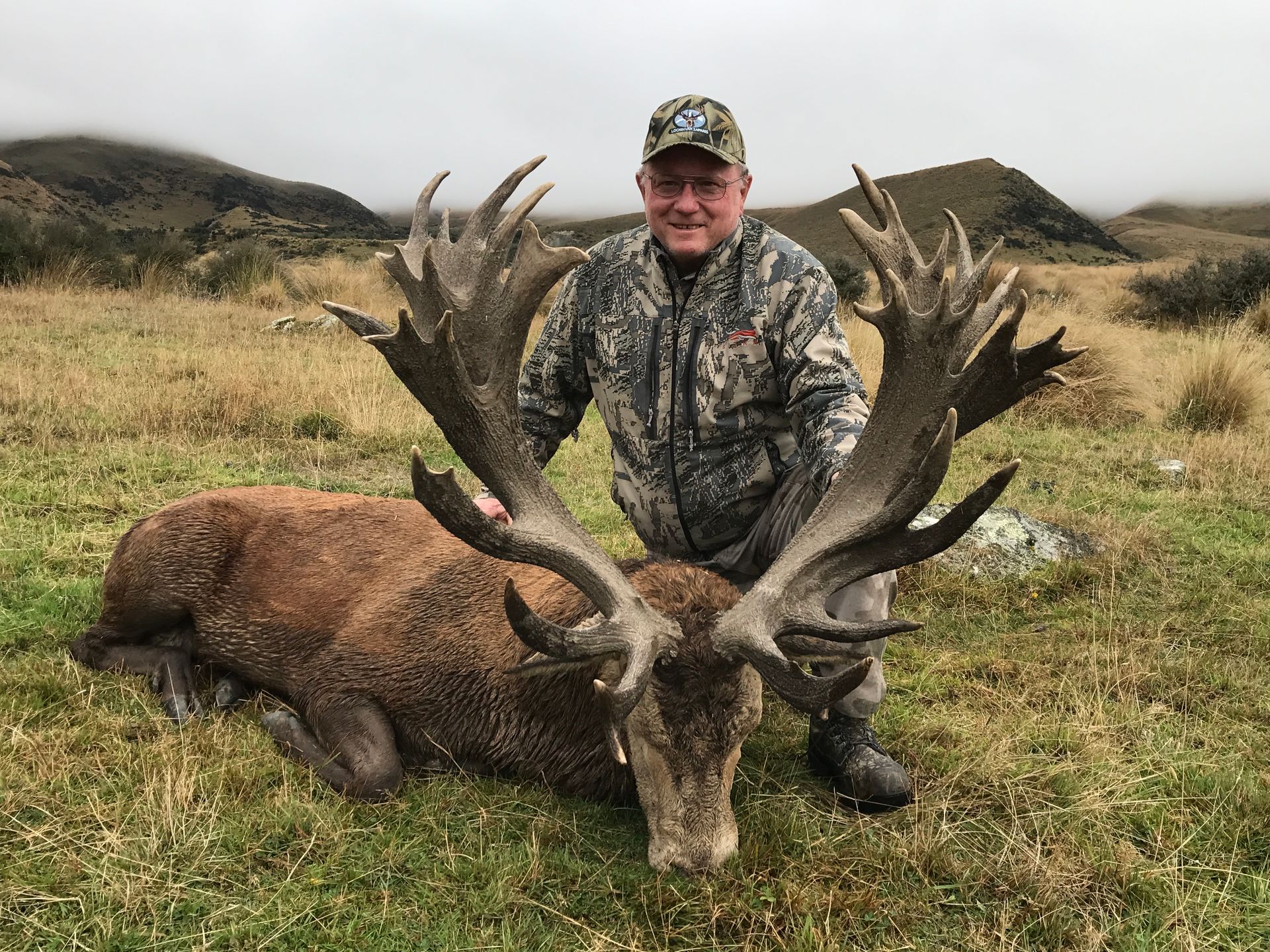 Man in camouflage poses with a large red deer he has hunted on a grassy hillside.