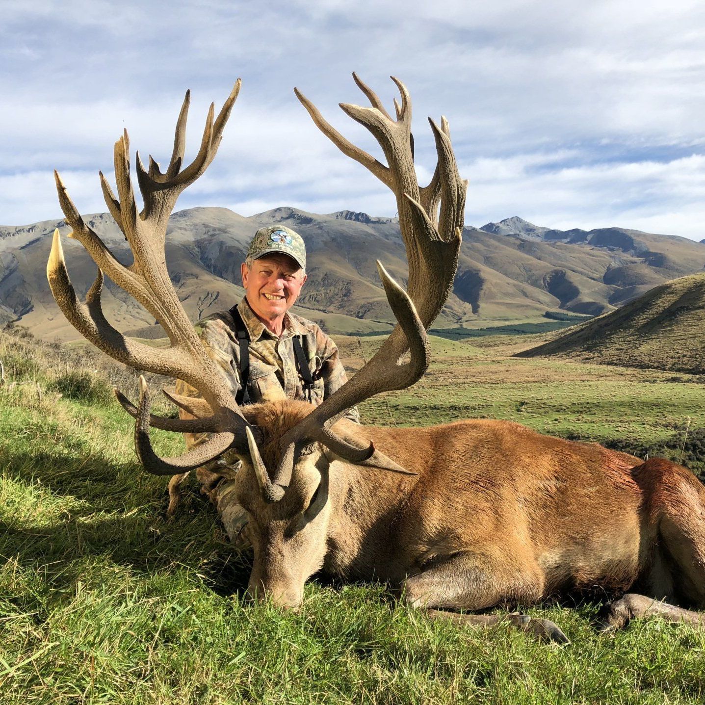 A man in camouflage smiling next to a large dead red deer with impressive antlers in a grassy, mountainous landscape.