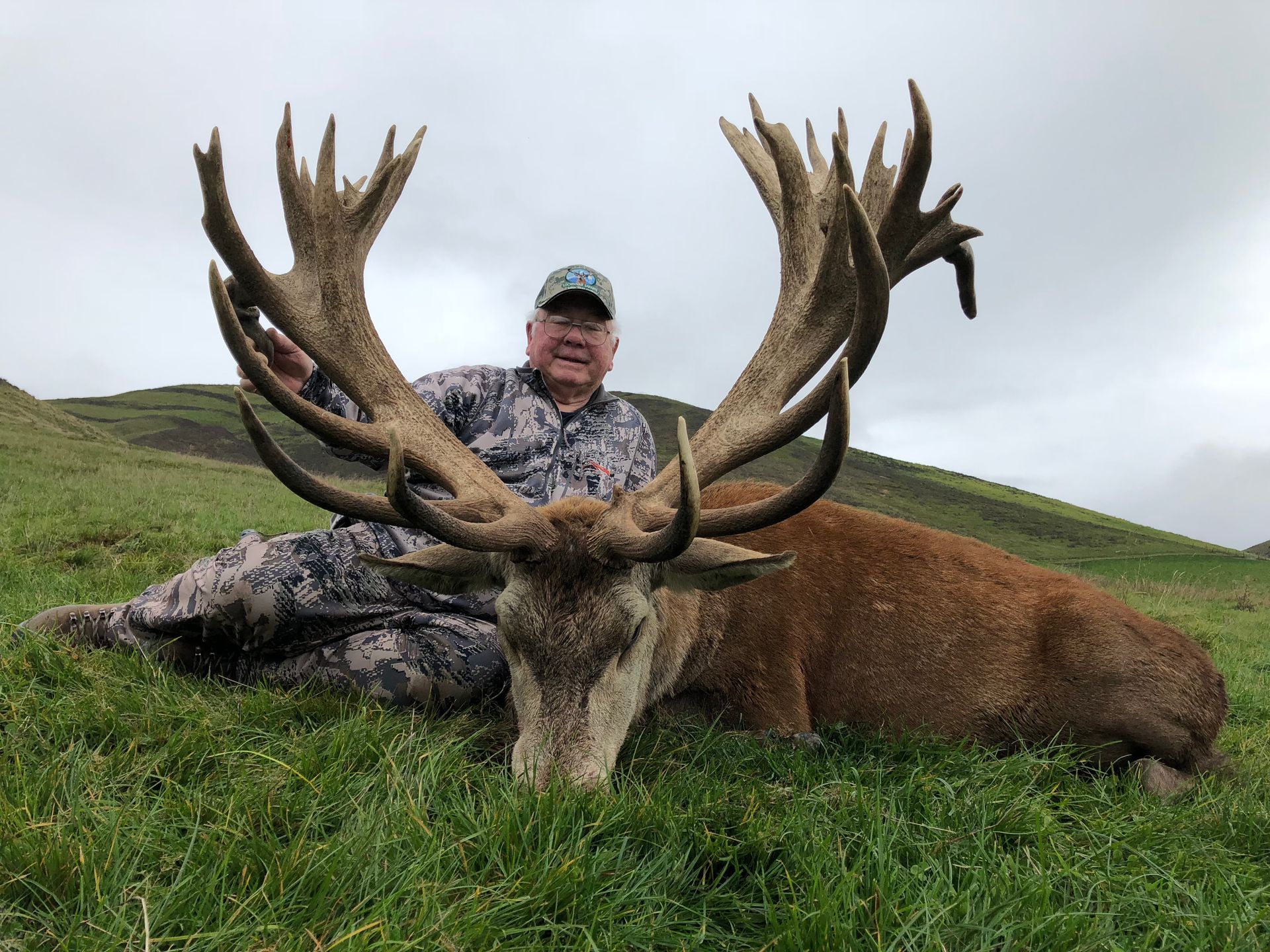 Man in camouflage rests by a large, dead red deer with impressive antlers on grassy terrain.