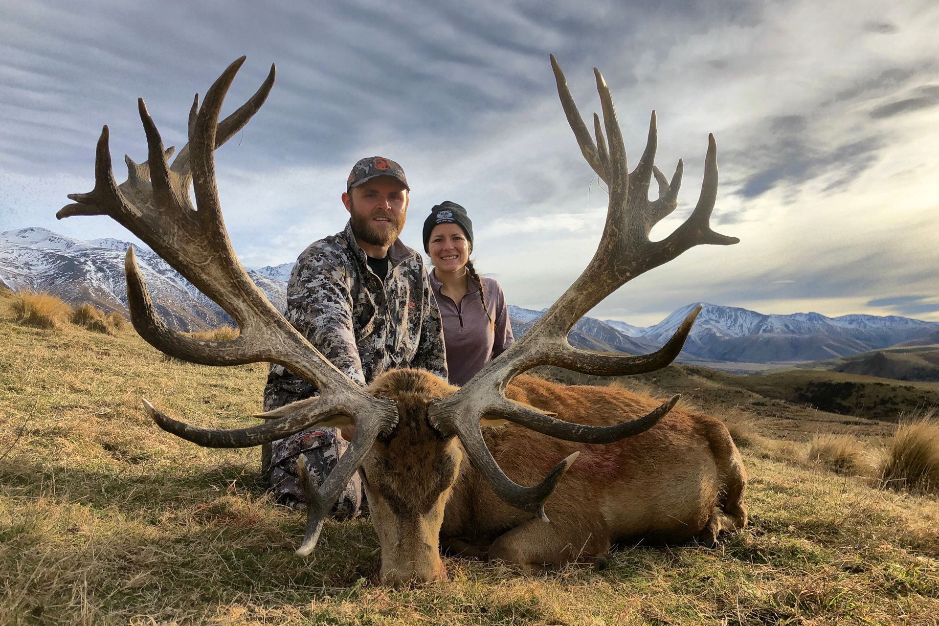 Two people stand next to a large dead deer with massive antlers. They are in a grassy mountain setting.