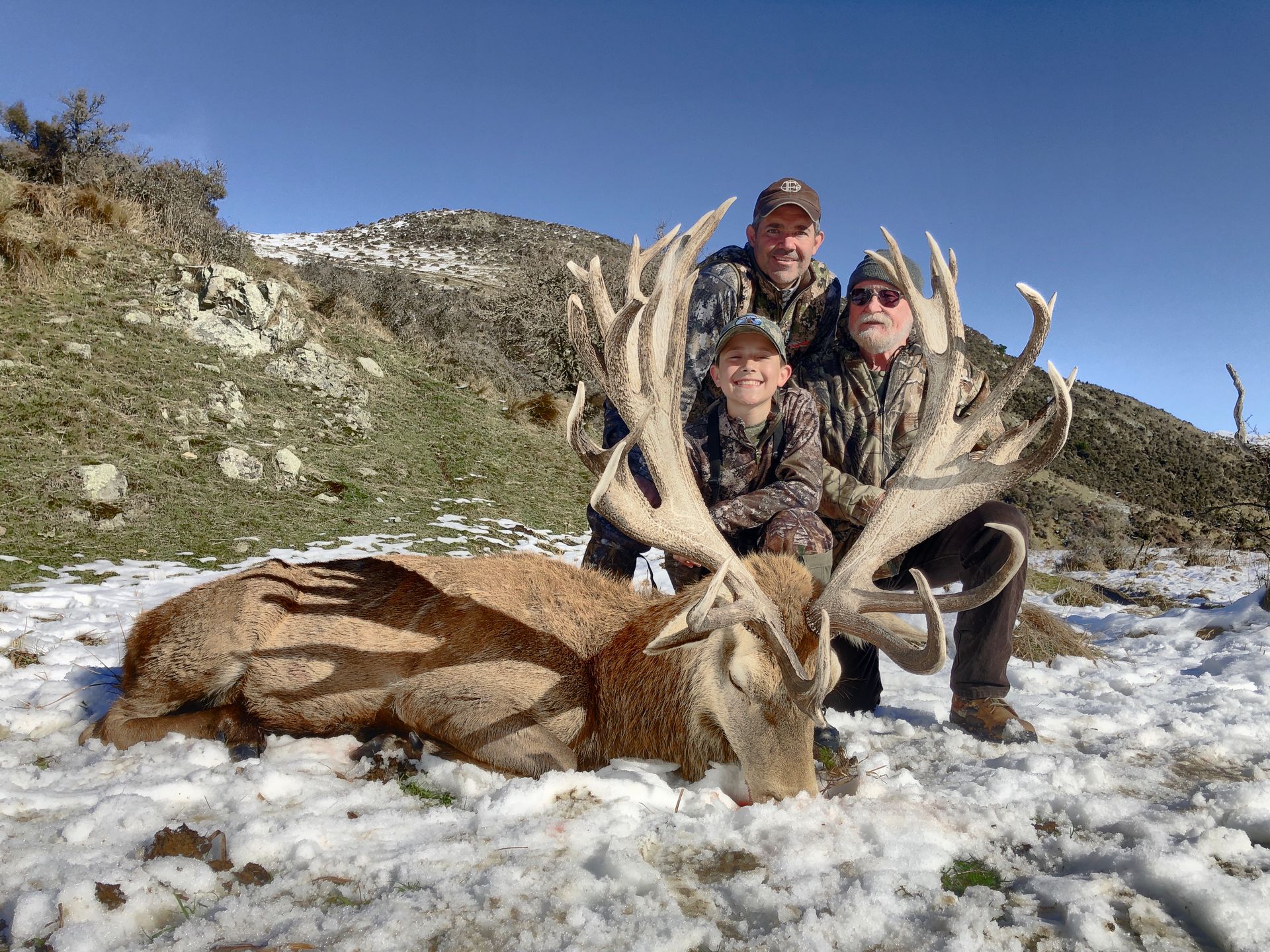 A family poses with a large red stag on snowy ground, mountains in the background. The stag has impressive antlers. All are smiling and in camouflage.