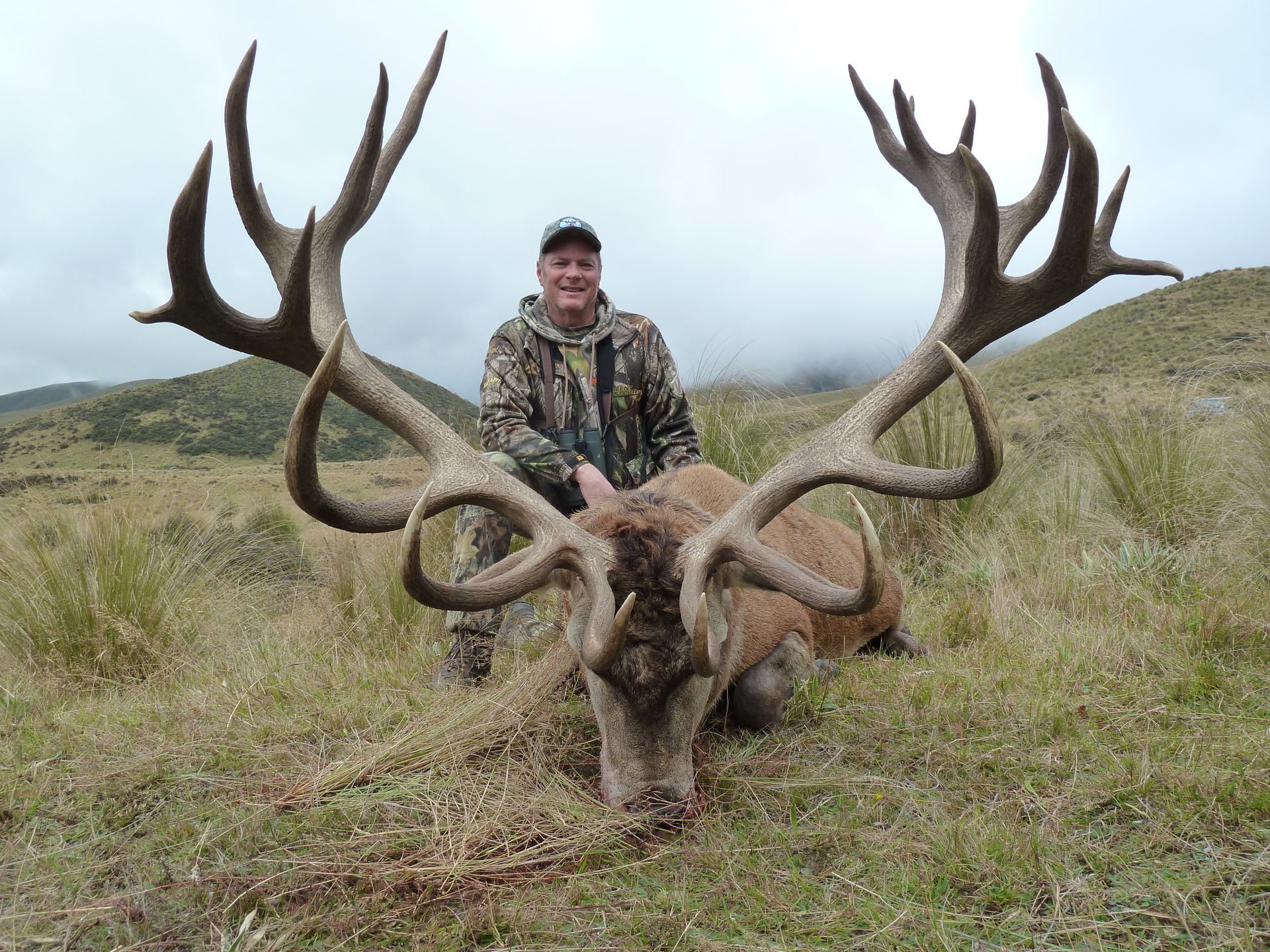 Man in camouflage standing with a large deer carcass and massive antlers in a grassy field.