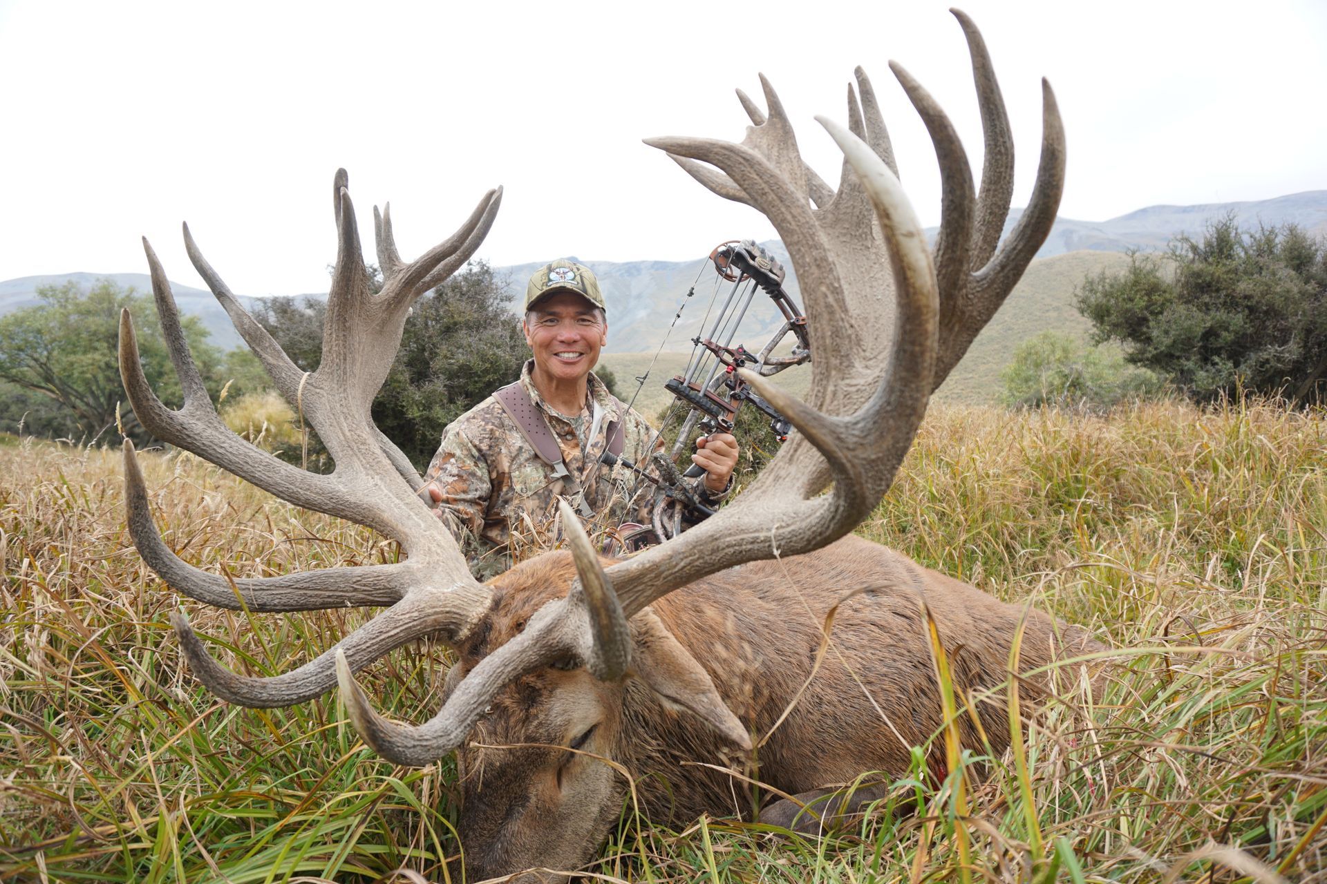 A smiling person in camouflage holding a bow next to a large dead stag with impressive antlers in a grassy field.