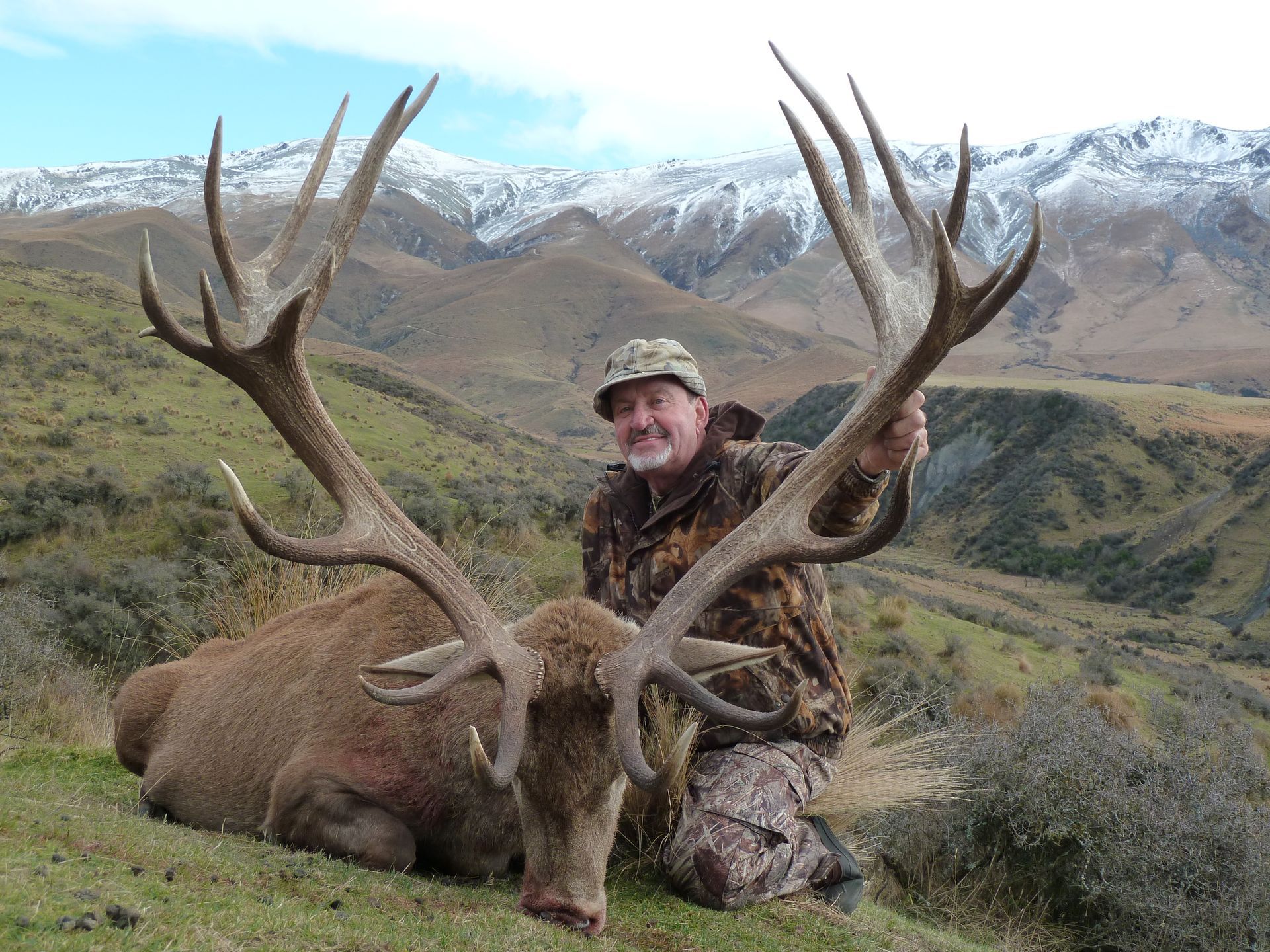Hunter kneels beside a large red deer with massive antlers in a mountainous landscape. The hunter smiles, wearing camouflage.