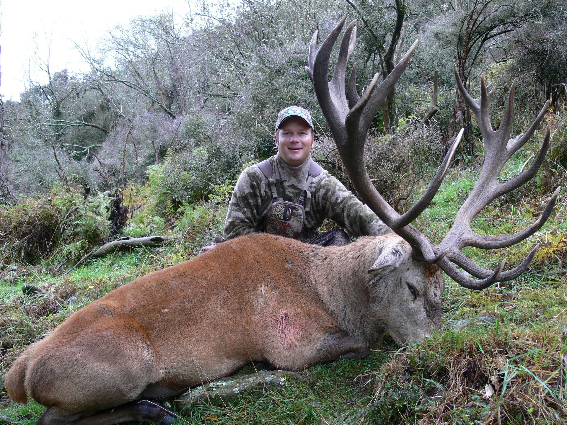 Hunter in camouflage kneels beside a large, reddish deer with impressive antlers on a grassy hillside.
