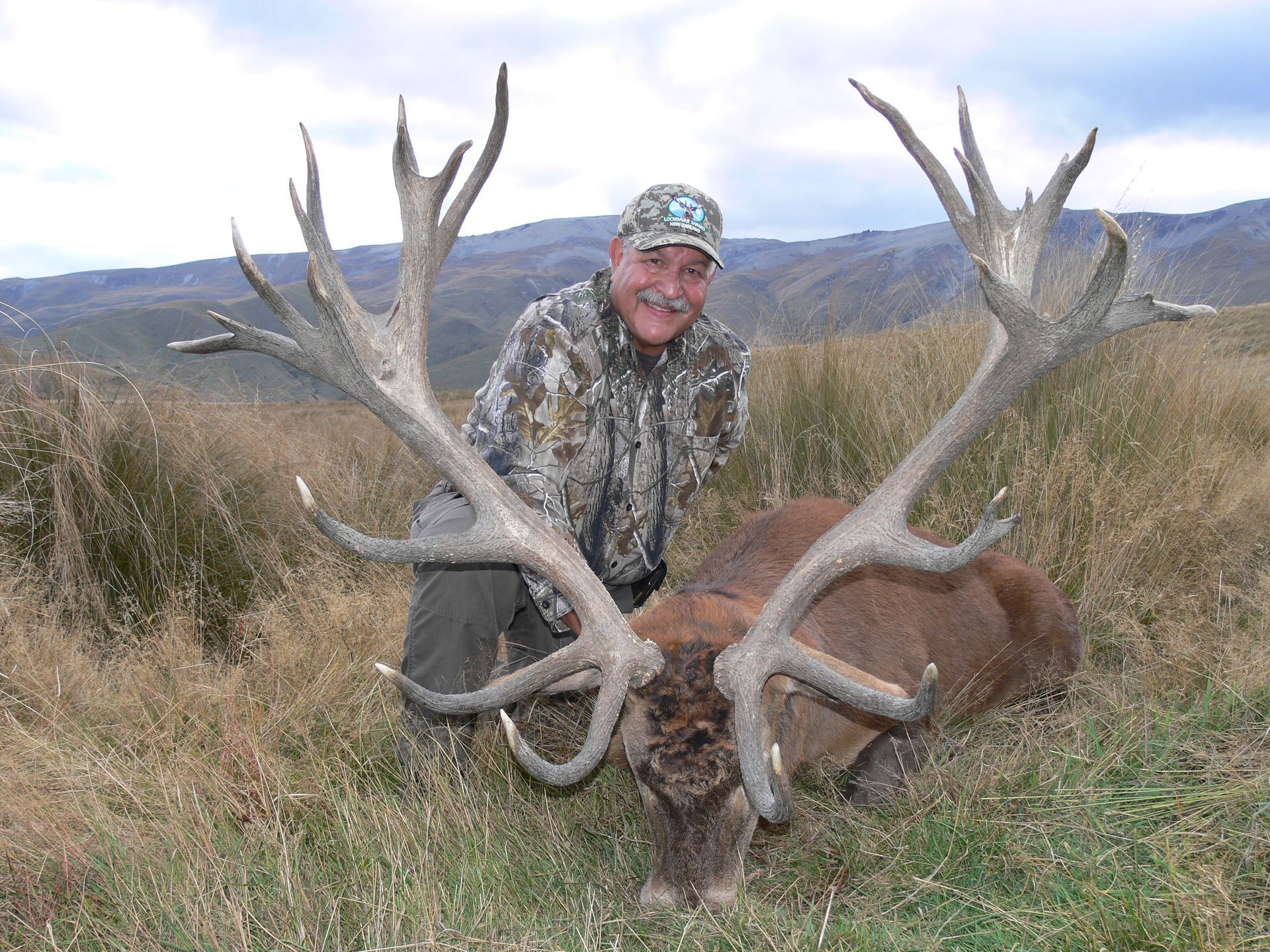 Man in camouflage kneeling beside a large red deer with impressive antlers, in a grassy, mountainous setting.