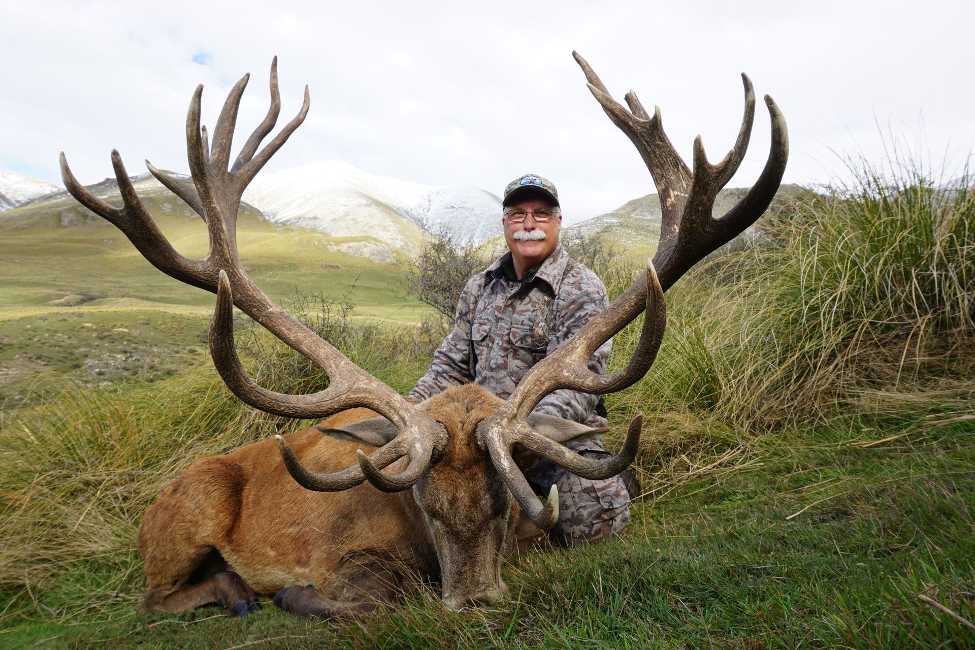 A man in camouflage kneels beside a large dead red deer with massive antlers in a grassy field, mountains in the background.