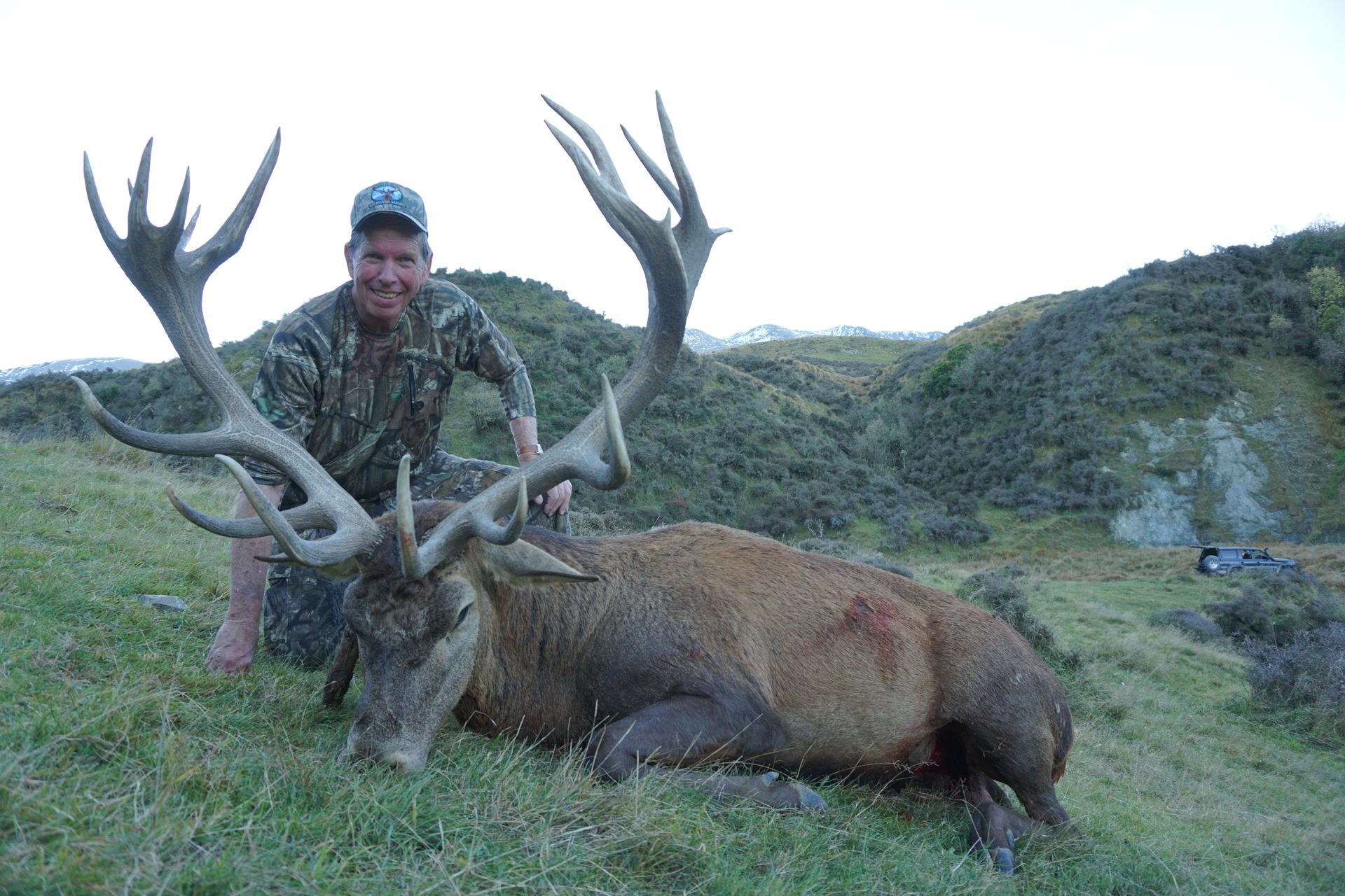 A man in camouflage kneels next to a large red deer stag with impressive antlers on a grassy hillside.