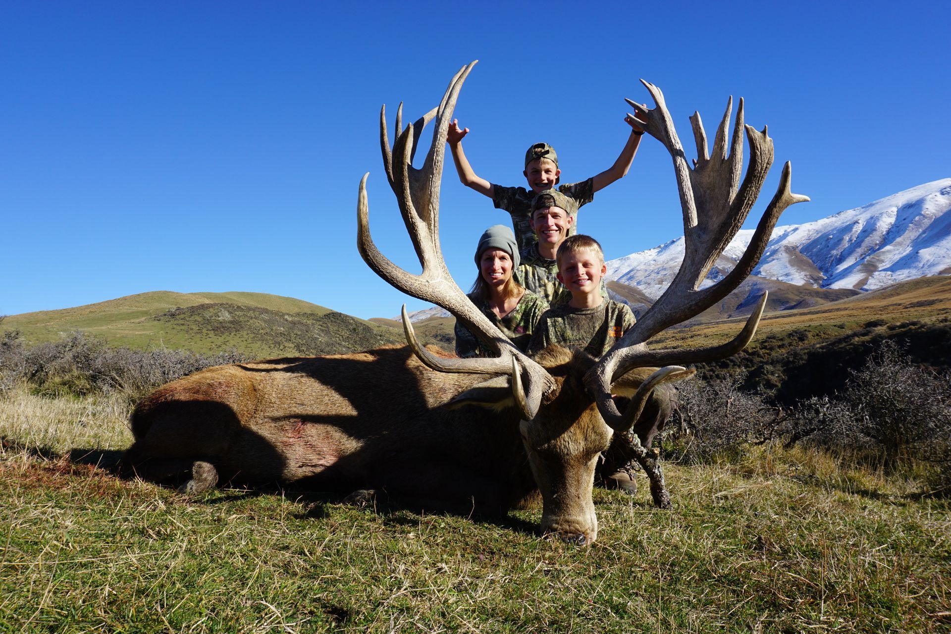 Three people posing with a large dead red deer in a grassy field, mountains in the background. The sky is clear and blue.