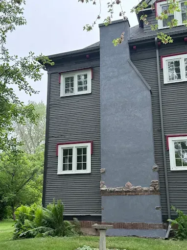 Gray-sided house with white-trimmed windows and a central stucco chimney, viewed from the front amid trees