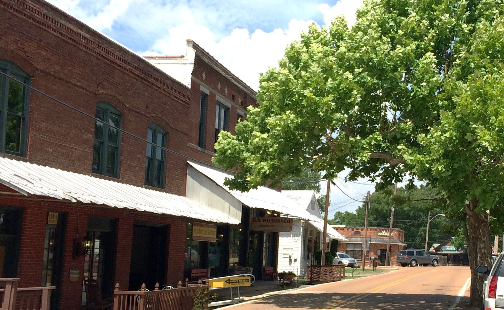 Historic brick storefronts along a quiet street with awnings and a large green tree on the sidewalk