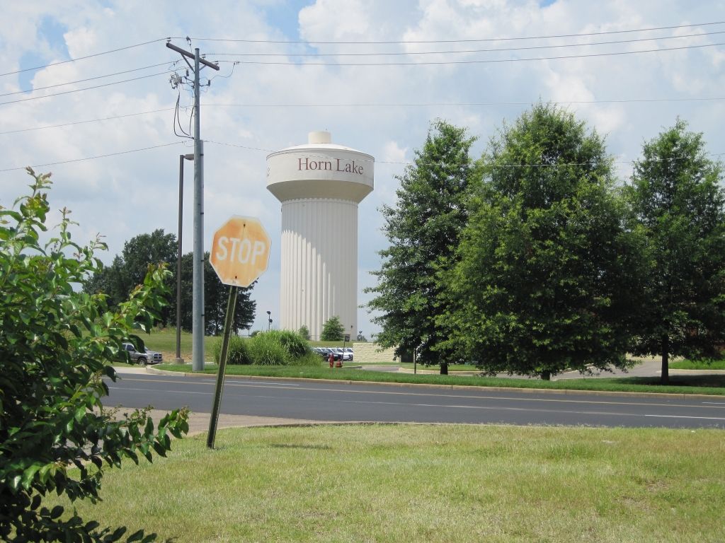Small-town street with a stop sign and a white water tower labeled “Huron Lake” under a cloudy sky