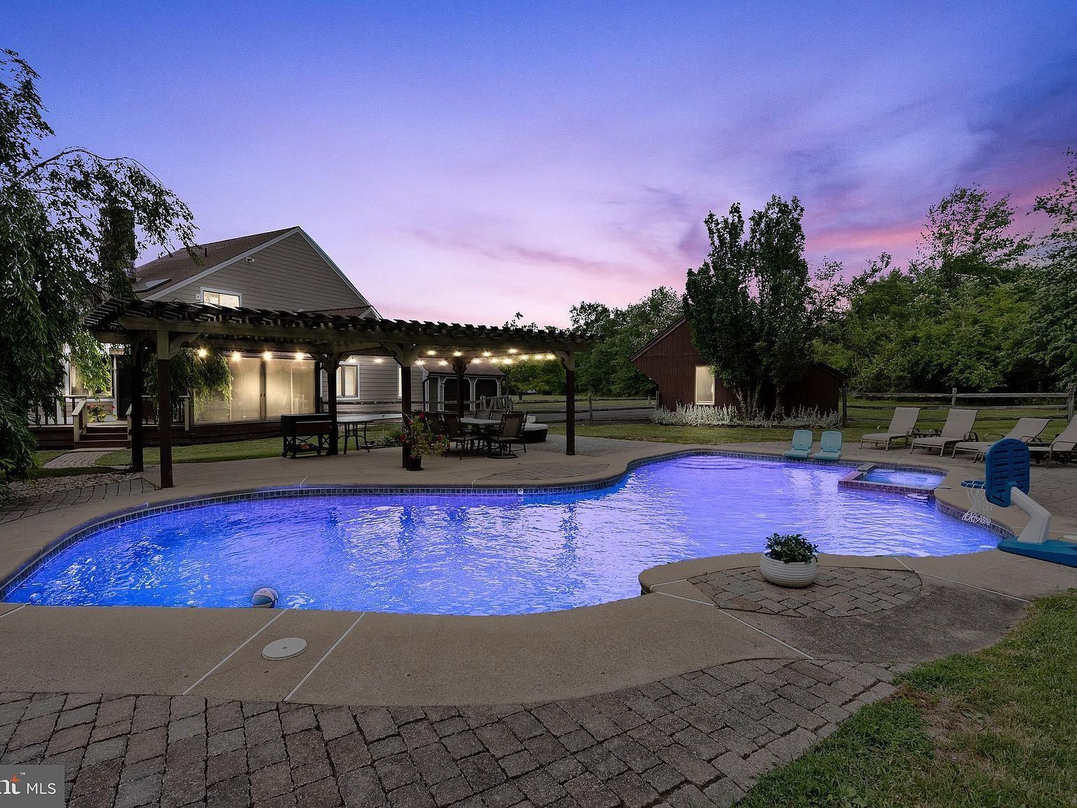 Backyard pool at dusk with blue-lit water, pavilion lights, and a pink-purple sky