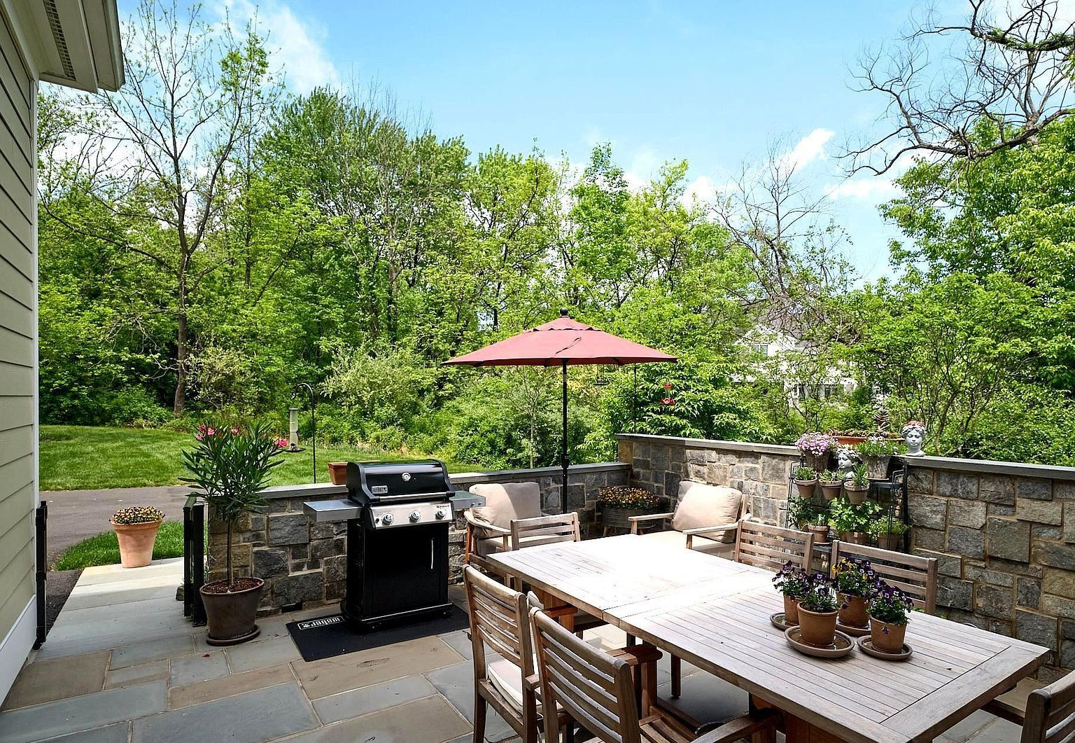 Patio with dining table, grill, umbrella, and potted plants overlooking a green backyard