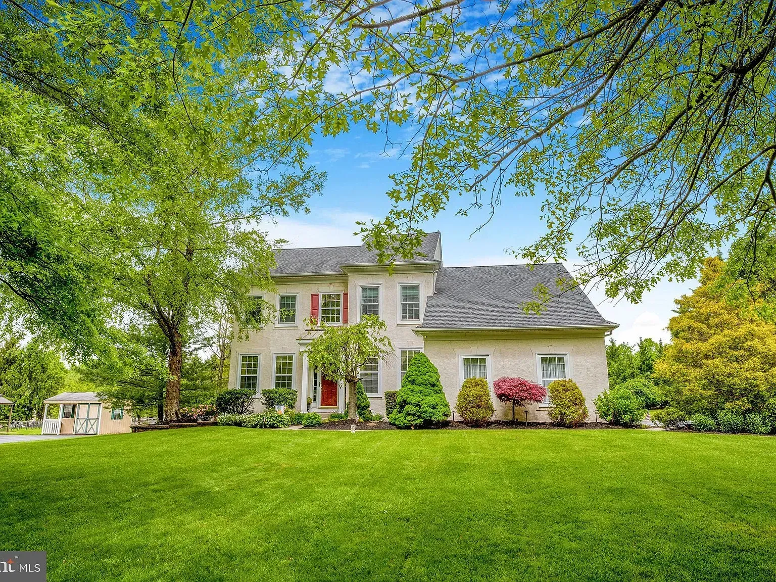 White two-story house with red front door, framed by lush green trees and lawn under a blue sky