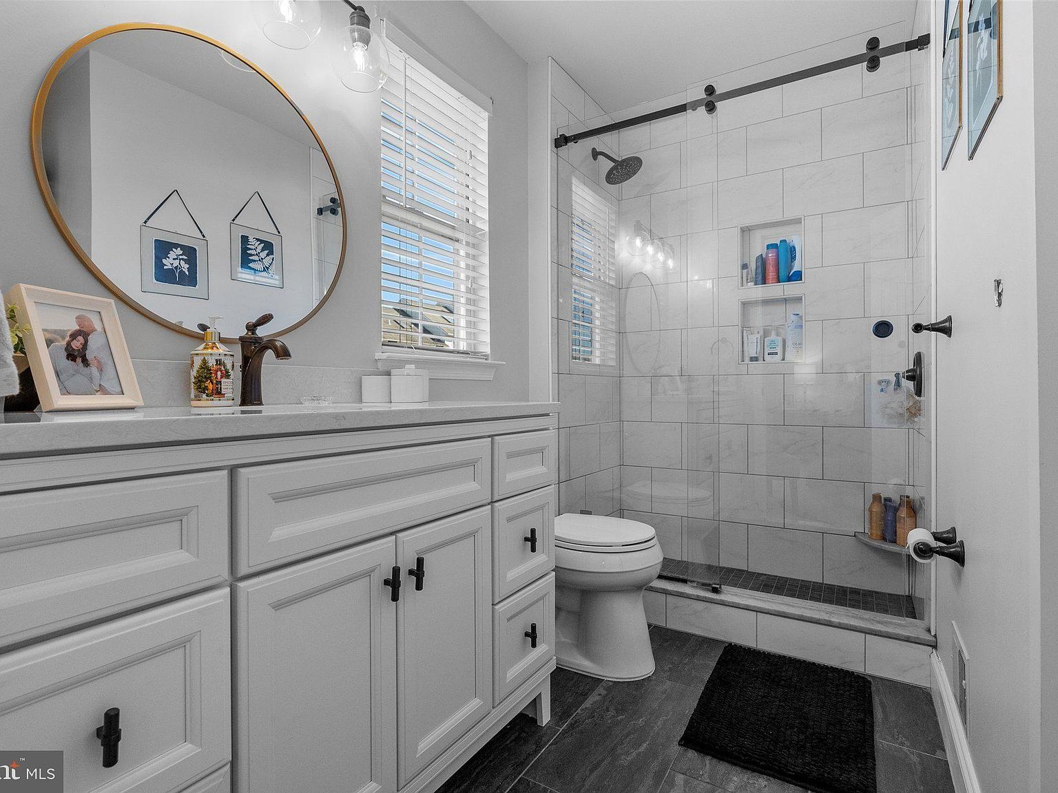Bright white bathroom with vanity, round mirror, shower, toilet, and black rug
