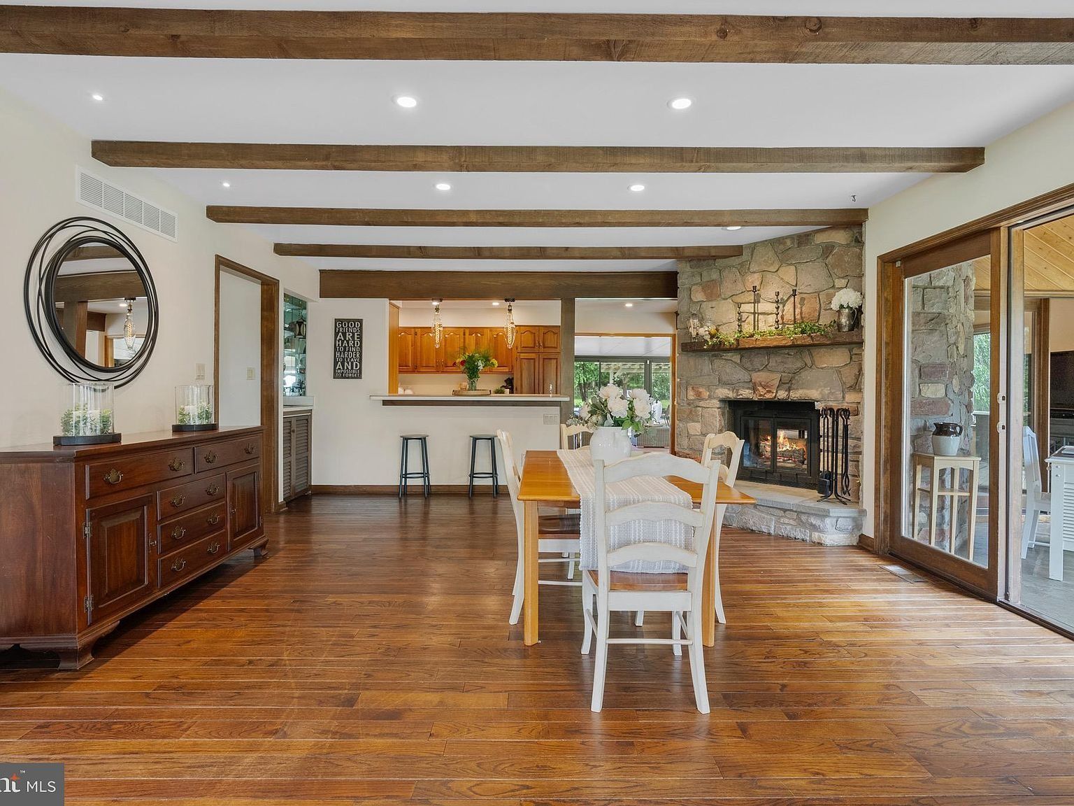 Open dining room with hardwood floors, wood beams, white table and chairs, and a stone fireplace.