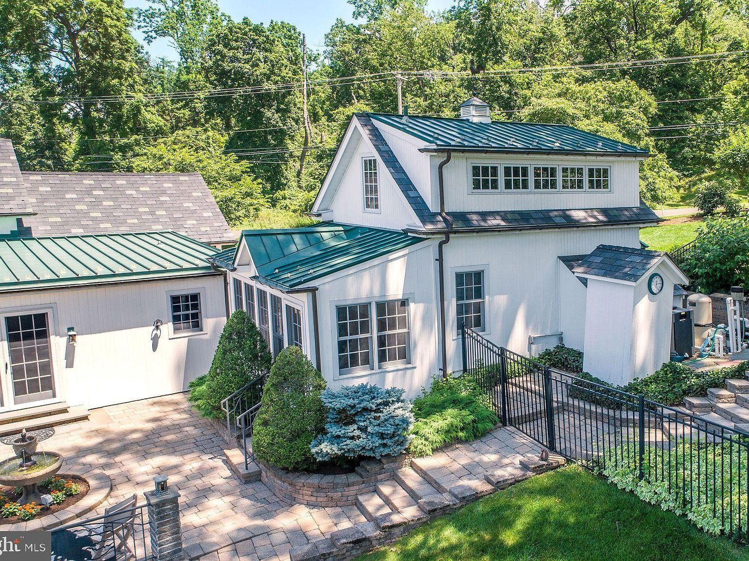 White house with green metal roofs, stone patio, and black fence in a leafy garden.