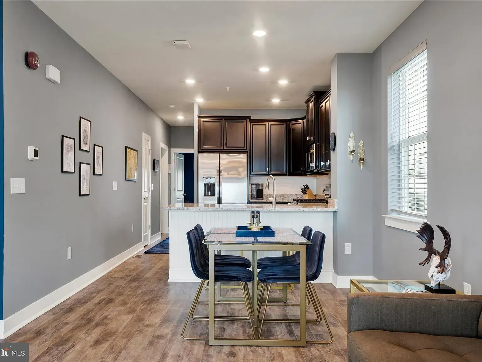 Open-concept living room and kitchen with gray walls, wood floors, blue rug, and dining table by a window.