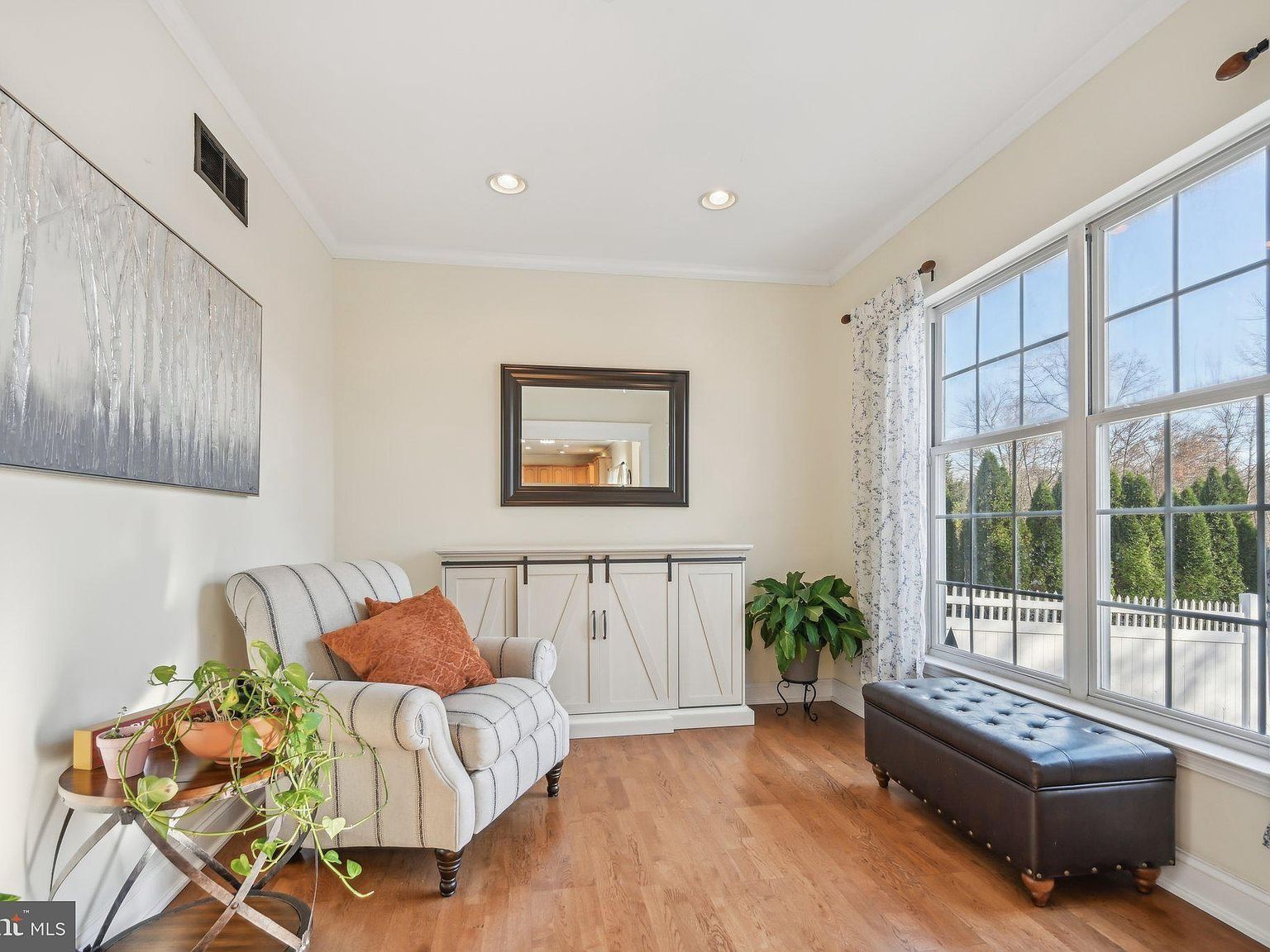Bright modern living room with striped armchair, white cabinet, mirror, plant, and brown ottoman by windows.