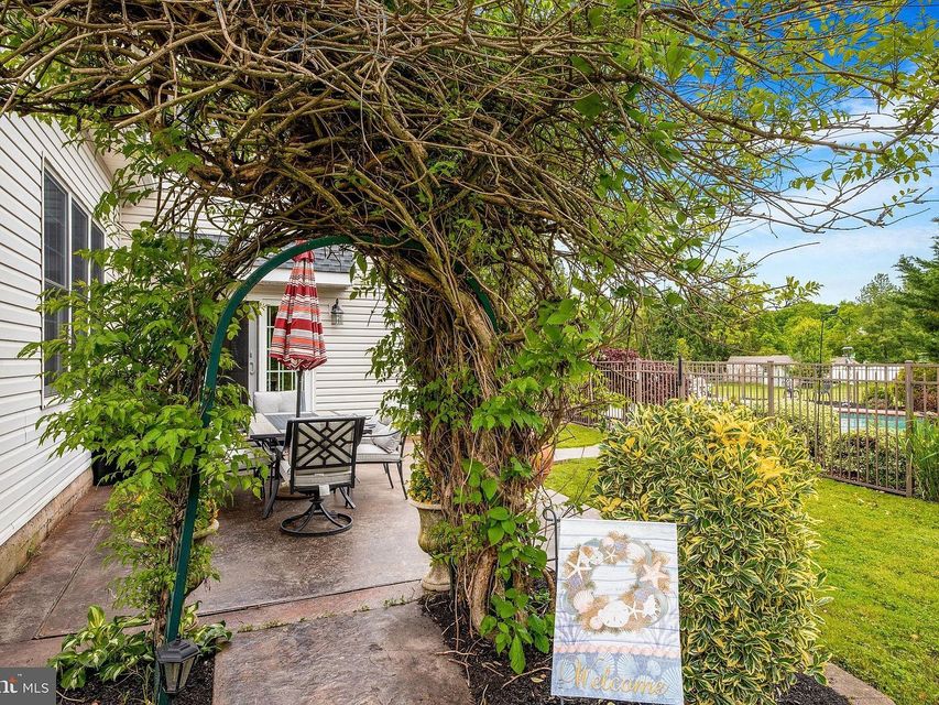 Vine-covered garden arch leading to a patio with table and chairs beside a white house