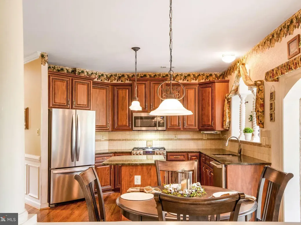 Warm kitchen with wooden cabinets, stainless fridge, and a round dining table by a sunlit window.