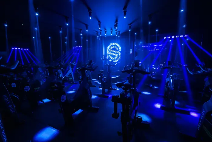 Indoorfietsstudio met blauwe verlichting, fietsers en een lichtgevend logo aan de muur
