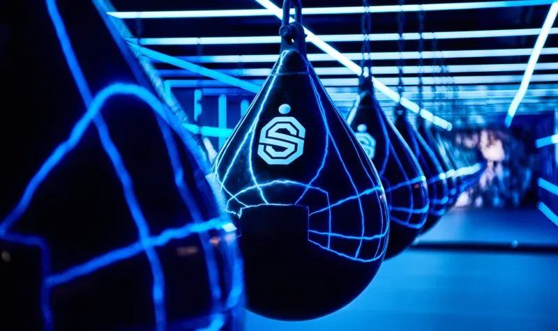 Blauwe bokszakken die oplichten in een neonverlichte sportschool, waarvan één tas is voorzien van een ‘S’-logo