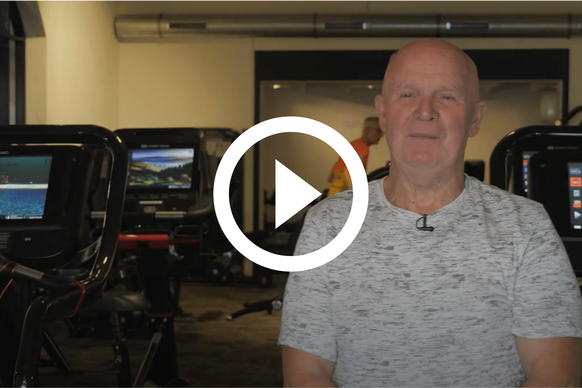 Man zit in een sportschool naast fitnessapparaten, met een afspeelknop voor video in beeld.