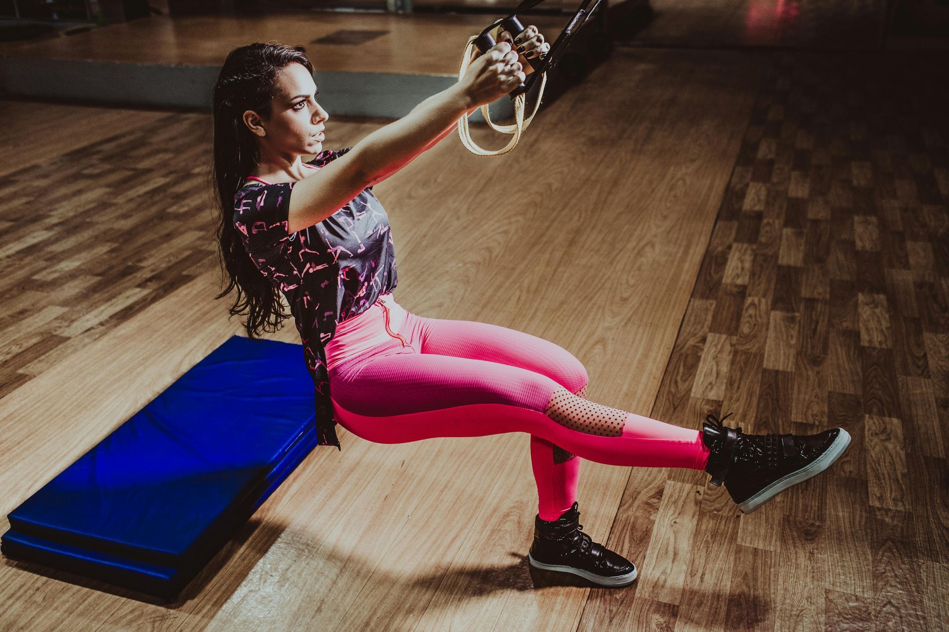 Persoon die zittend een kettlebelloefening doet in een sportschool, met een roze legging naast een blauwe mat.