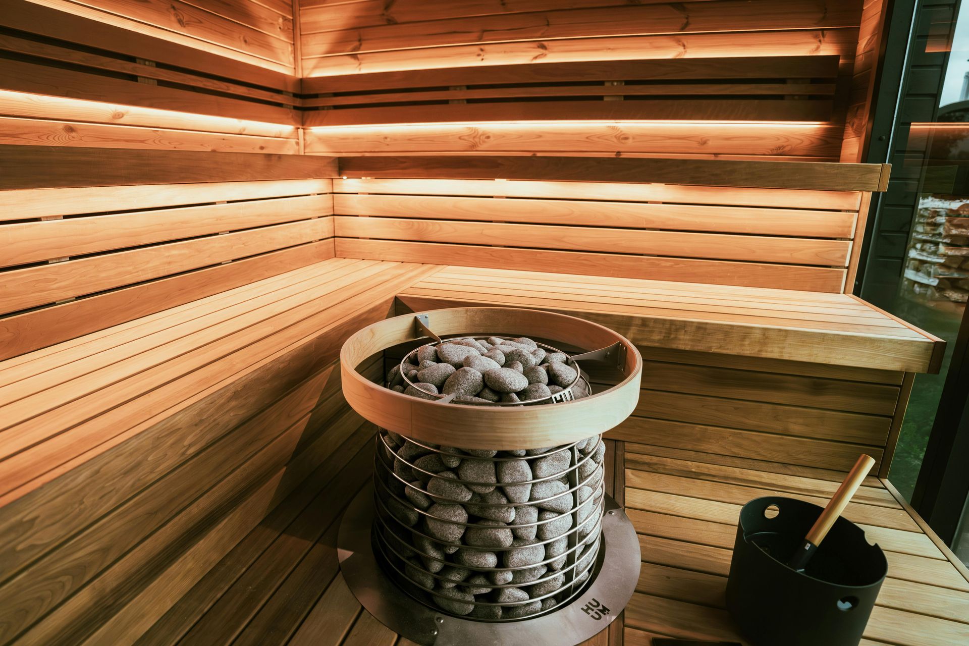 Sauna met houten wandbekleding, een ronde stenen kachel en sfeervolle verlichting, met een emmer en een houten lepel.