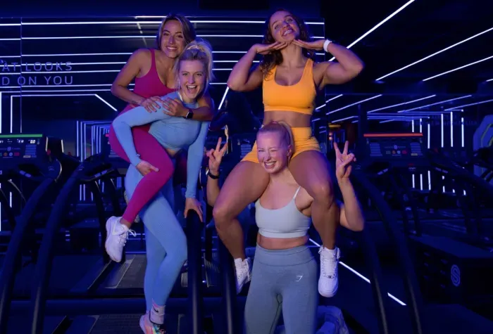 Vier vrouwen poseren samen in een sportschool met neonverlichting, glimlachend en met vredestekens na een training.