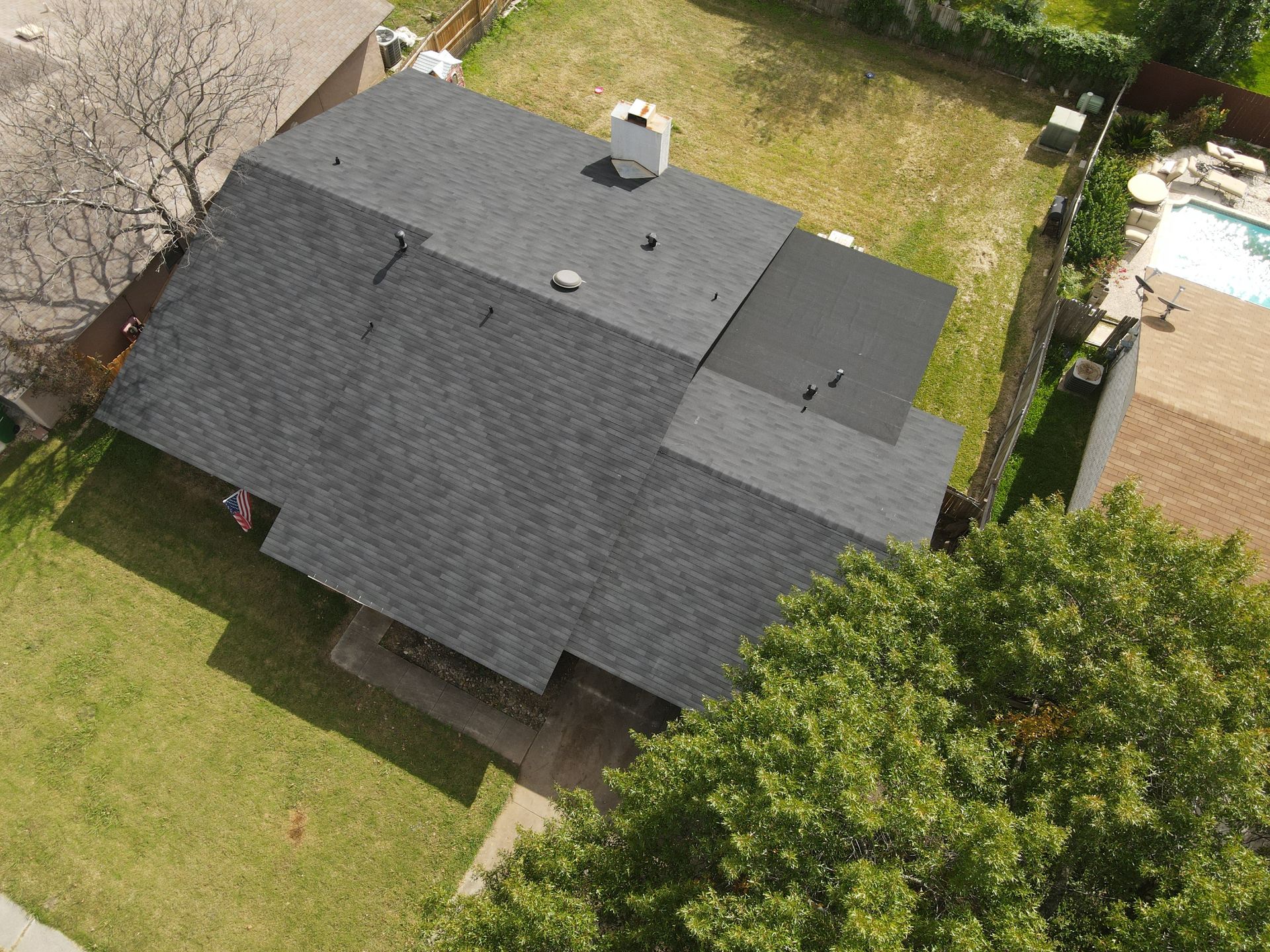 Overhead view of a house with a dark roof, surrounded by a green lawn and trees, sunny day.