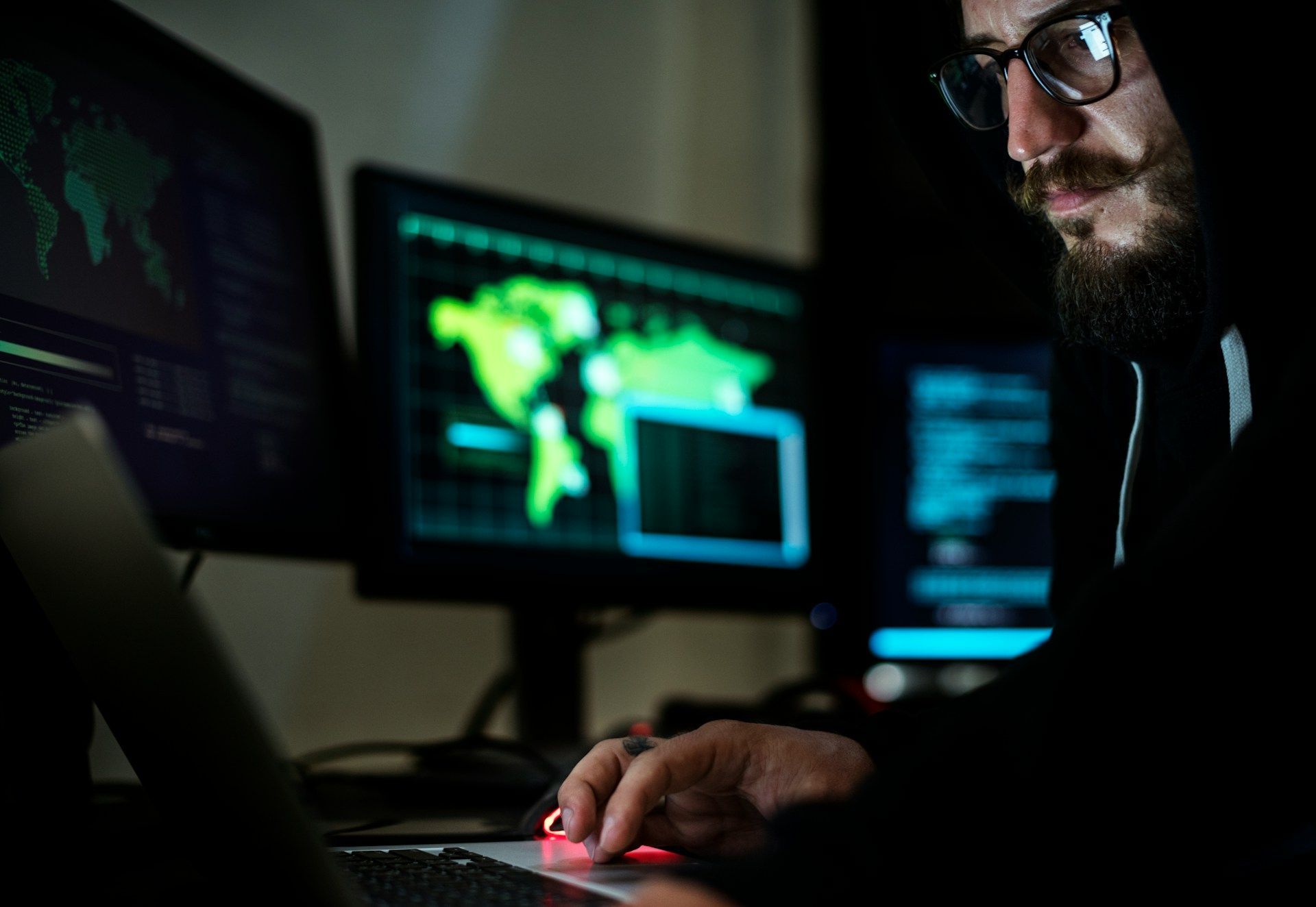 A man in a hood is using a laptop computer in front of two computer monitors.